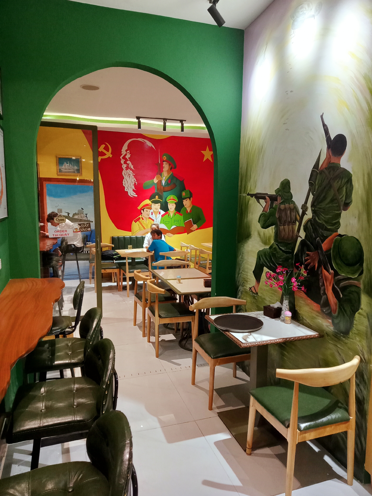 This image captures the interior of a cafe or restaurant in Nha Trang, Vietnam, distinguished by its strong thematic decor. The space is largely painted green, with a prominent arched doorway dividing the area.
On the left, beyond the archway, a vivid red wall features a large mural. This mural prominently displays the hammer and sickle symbol, an iconic depiction of Ho Chi Minh, and several figures in green military uniforms with red hats, suggesting a scene of unity or celebration. In this section, a person in a white shirt is visible standing at a counter, possibly placing an order. Another person in a blue shirt is seated at a table with dark green upholstered booths. A framed painting of a sailing ship hangs on a yellow wall segment. Text visible on a sign near the counter reads "Open" and "ORDER TẠI QUẦY" (Order at counter).
The main seating area, in the foreground and to the right, features a large mural on a green-painted wall. This mural depicts several soldiers in green military uniforms, armed with rifles and an RPG launcher, positioned as if in a jungle or forest environment with light filtering through trees, evoking a scene of wartime or struggle. The area is furnished with light wooden tables and chairs, which feature dark green leatherette seats. Some tables are accompanied by dark green upholstered booths. On a table in the foreground, a vase with pink flowers and a tissue box are placed. Overhead, track lighting and recessed lights illuminate the indoor setting. The imagery and military themes strongly suggest a cafe with a Vietnamese historical or political motif.