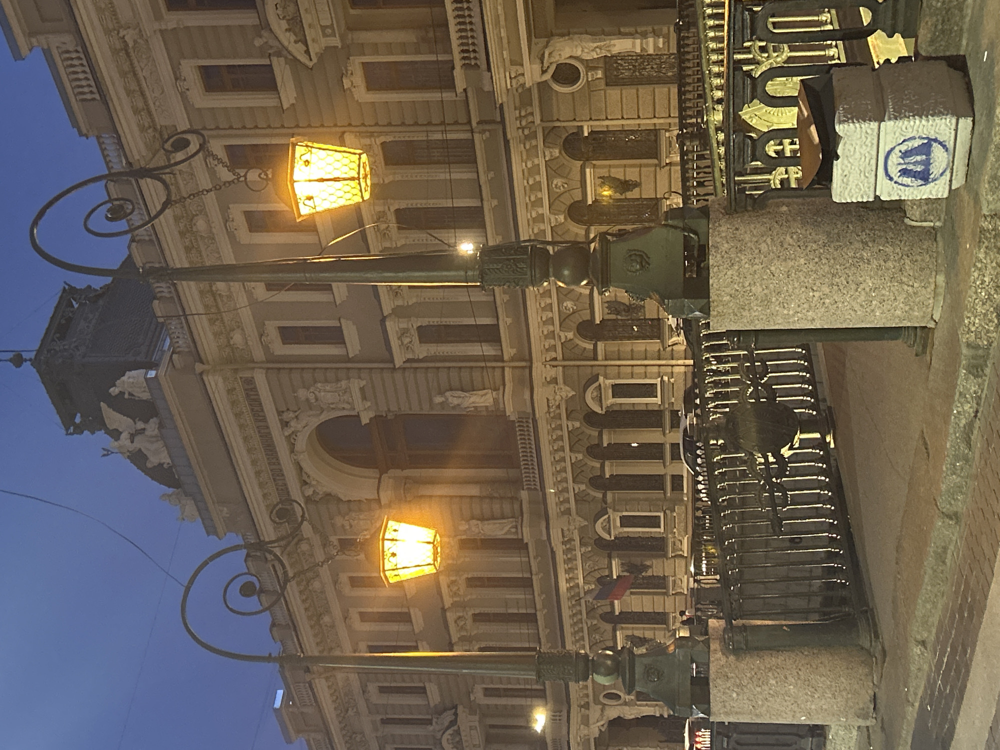The photograph captures a building facade at dusk, illuminated by decorative streetlights. Two ornate lampposts with yellow-tinted lanterns stand in the foreground, flanking a metal railing that appears to be part of a bridge or embankment. The building behind them is multi-story, with classical architectural details, including columns, arched windows, and sculpted figures. The sky is a deep blue, suggesting early evening. The street in front of the railing is paved with cobblestones. To the right of the frame, on the ground, is a concrete block with a blue symbol resembling three arched structures.