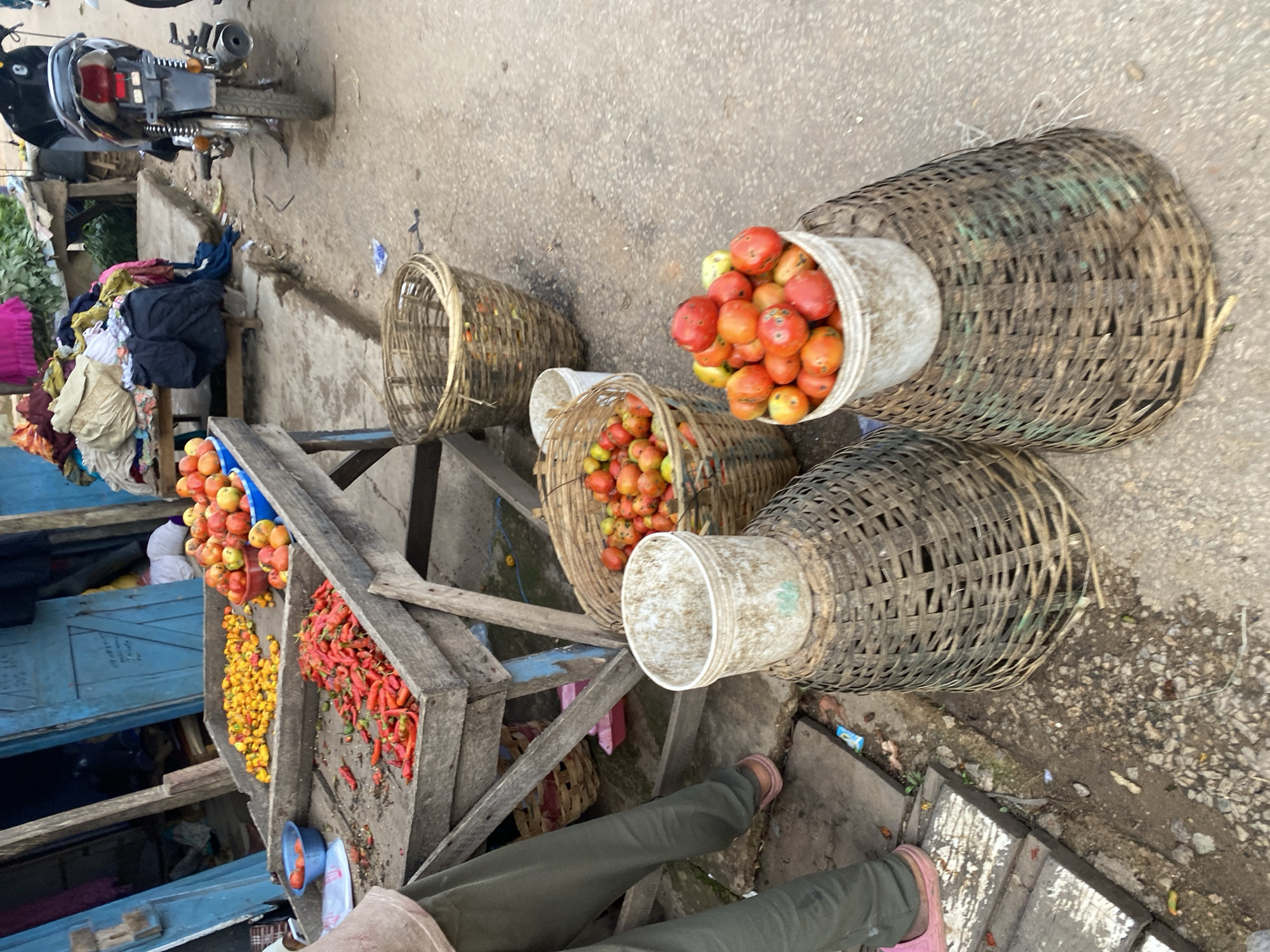 The image depicts a bustling outdoor market scene in Jos, Nigeria. In the foreground, a person, partially visible from the waist down, stands wearing olive green trousers and pink sandals. Their presence suggests they are either a vendor or a shopper.
Dominating the scene are various stalls laden with fresh produce. One stall, made of weathered wood, displays a vibrant assortment of red chili peppers. Adjacent to this, another section of the stall holds a pile of small, yellow peppers. Further back, a blue basin overflows with ripe tomatoes.
Several woven baskets are strategically placed around the produce. A large, dark woven basket sits in the middle ground, filled with a variety of red and green tomatoes. To the right, a similar, but smaller, woven basket also contains a collection of tomatoes. In the lower right corner, two large, conical woven baskets stand side-by-side, one supporting a white plastic bucket brimming with red tomatoes. Another white bucket, placed atop a woven basket, is visible behind the stall with chili peppers.
In the background, a black motorcycle is parked, hinting at the modes of transportation common in the area. Colorful fabrics and clothing items are draped over a nearby stall, adding to the vibrant market atmosphere. The ground is paved with concrete and dirt, indicative of an urban street environment. The overall impression is one of active commerce and the lively presentation of fresh goods in a Nigerian marketplace.