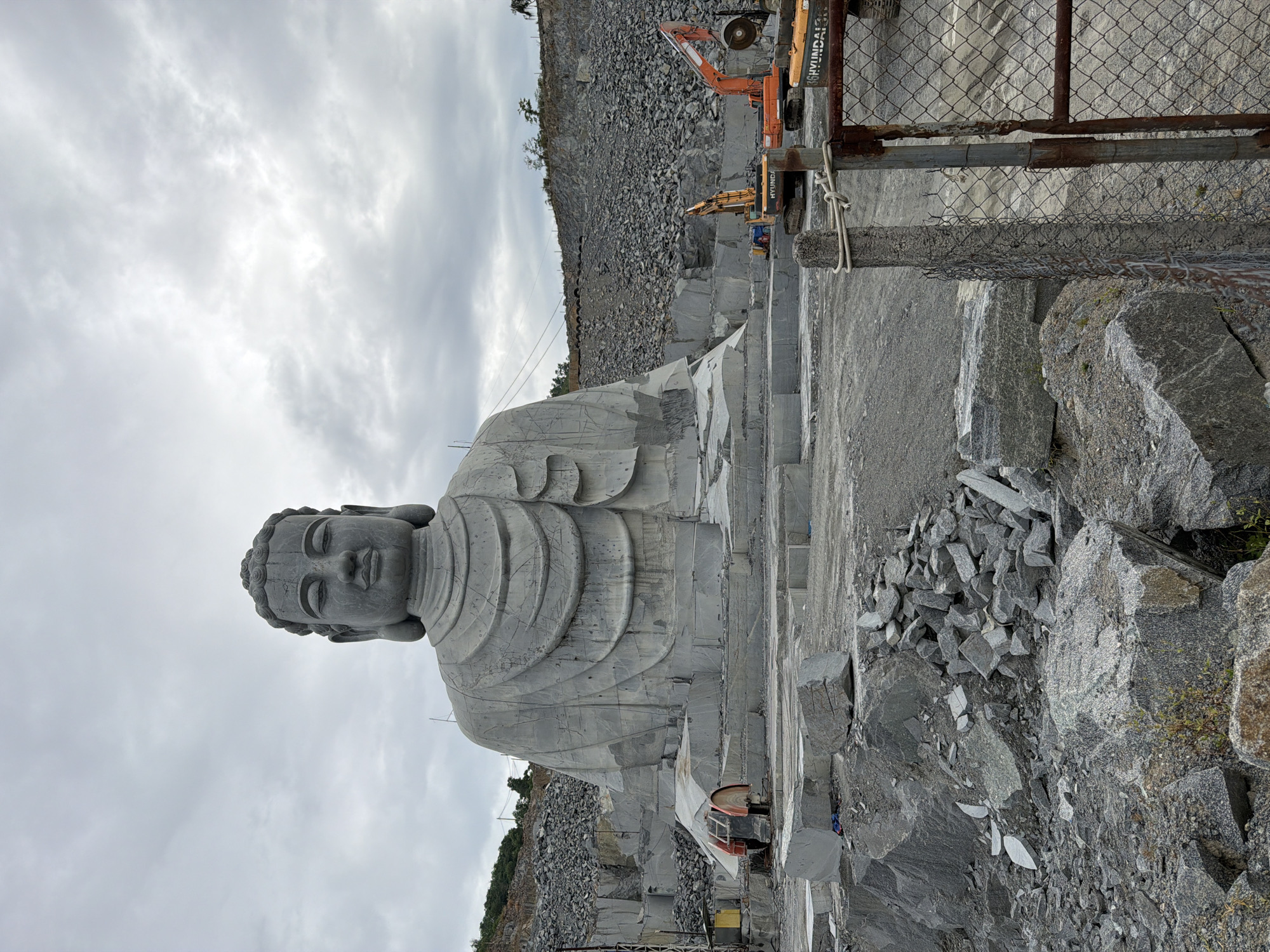 The image shows a large stone Buddha statue in a quarry-like setting. The statue appears to be unfinished or in the process of being carved directly from the stone. The statue depicts a seated Buddha with a serene expression. The surrounding area is filled with rocks, stones, and equipment, suggesting an active quarry or carving site. Heavy machinery like excavators and saws are visible in the background, along with a chain-link fence. The sky is overcast. The statue is located in Đà Nẵng, Vietnam.