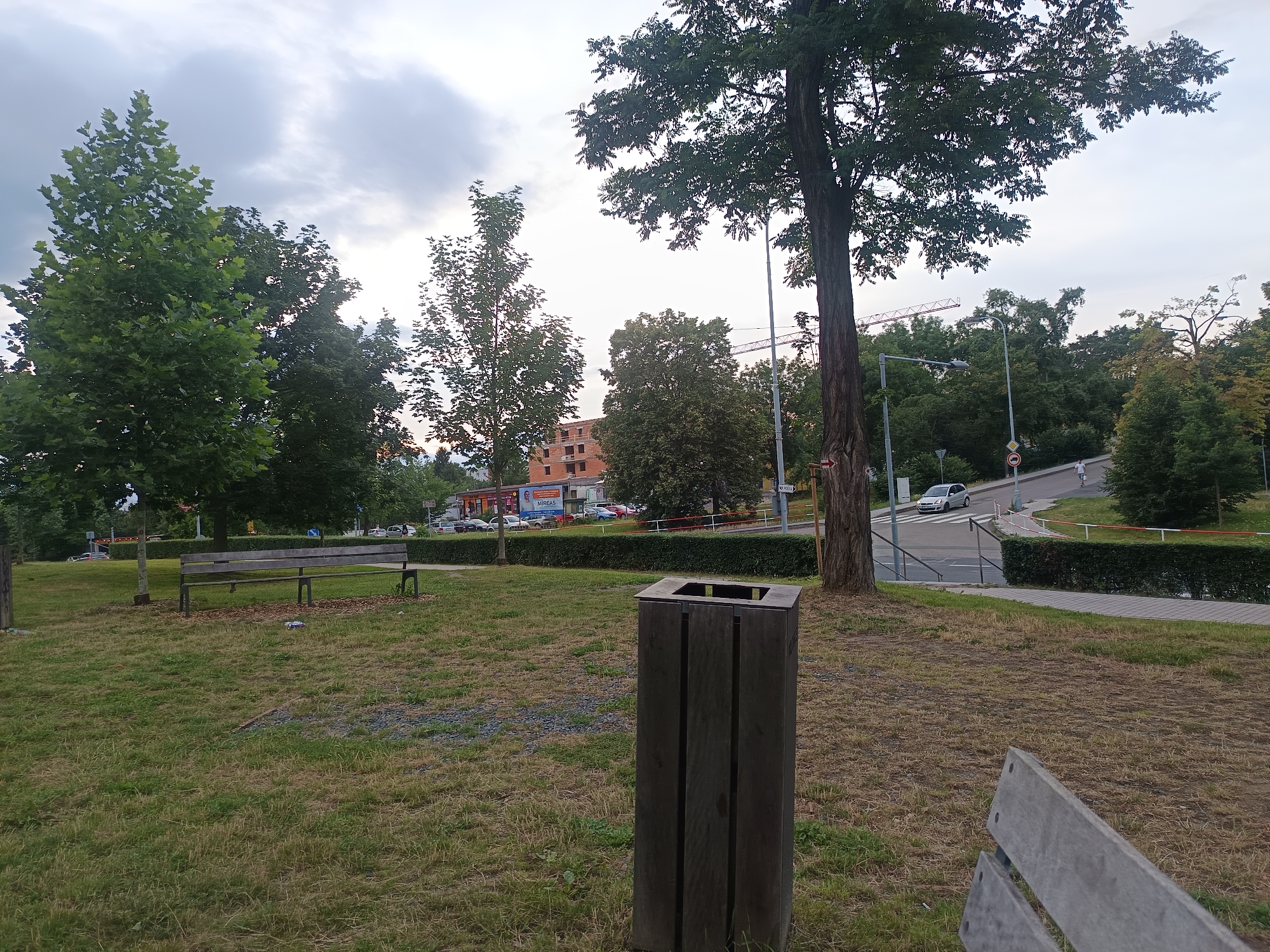 The image depicts an outdoor scene in the city of Milovice, Czechia, likely a park or public area. In the foreground, a weathered wooden trash receptacle stands prominently on dry grass. To the left, a wooden park bench is partially visible, also situated on the grass. The middle ground features a neatly trimmed hedge bordering a paved walkway. Beyond the hedge, a street curves uphill, with a light-colored car driving on it. In the background, several trees with lush green foliage are present. Across the street, a building under construction, with exposed brickwork, is visible. Red construction cranes are also visible in the distance, indicating ongoing development. The sky is overcast with a mix of grey and white clouds.