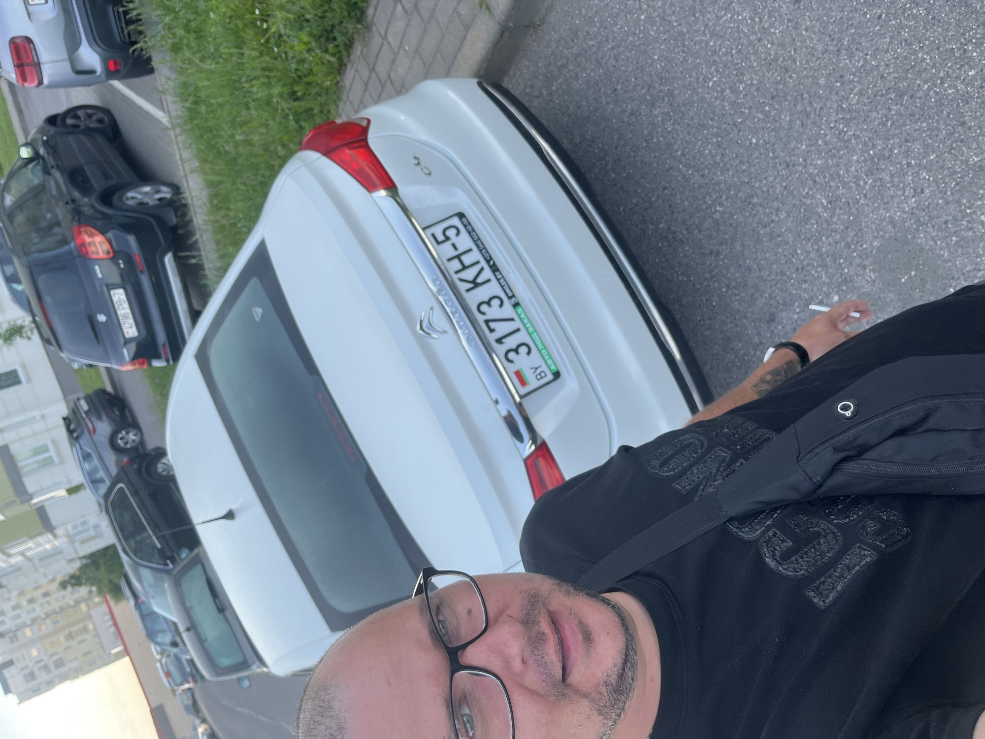 A man is captured in a close-up, selfie-style shot, looking directly at the camera with a neutral expression. He wears black-rimmed glasses, a black t-shirt with embossed text (appearing to be "ONE" and "LOVE"), and a black fanny pack or sling bag. He has a watch on his left wrist and holds a lit cigarette in his right hand.
The man is standing in a parking area in Minsk, Belarus. Behind him is a white Citroën C4, showing its rear with the model "C4" and brand "CITROËN" visible. Its Belarusian license plate reads "BY 3173 KH-5". The license plate frame also displays "АВТО ПОД ЗАКАЗ SMILES.BY" and a phone number "+375 (44) 552-55-00". Further back, a black Suzuki Swift and other cars are parked on asphalt next to a strip of green grass. Residential buildings can be seen in the distant background. The lighting suggests it is daytime, possibly late afternoon or early evening.