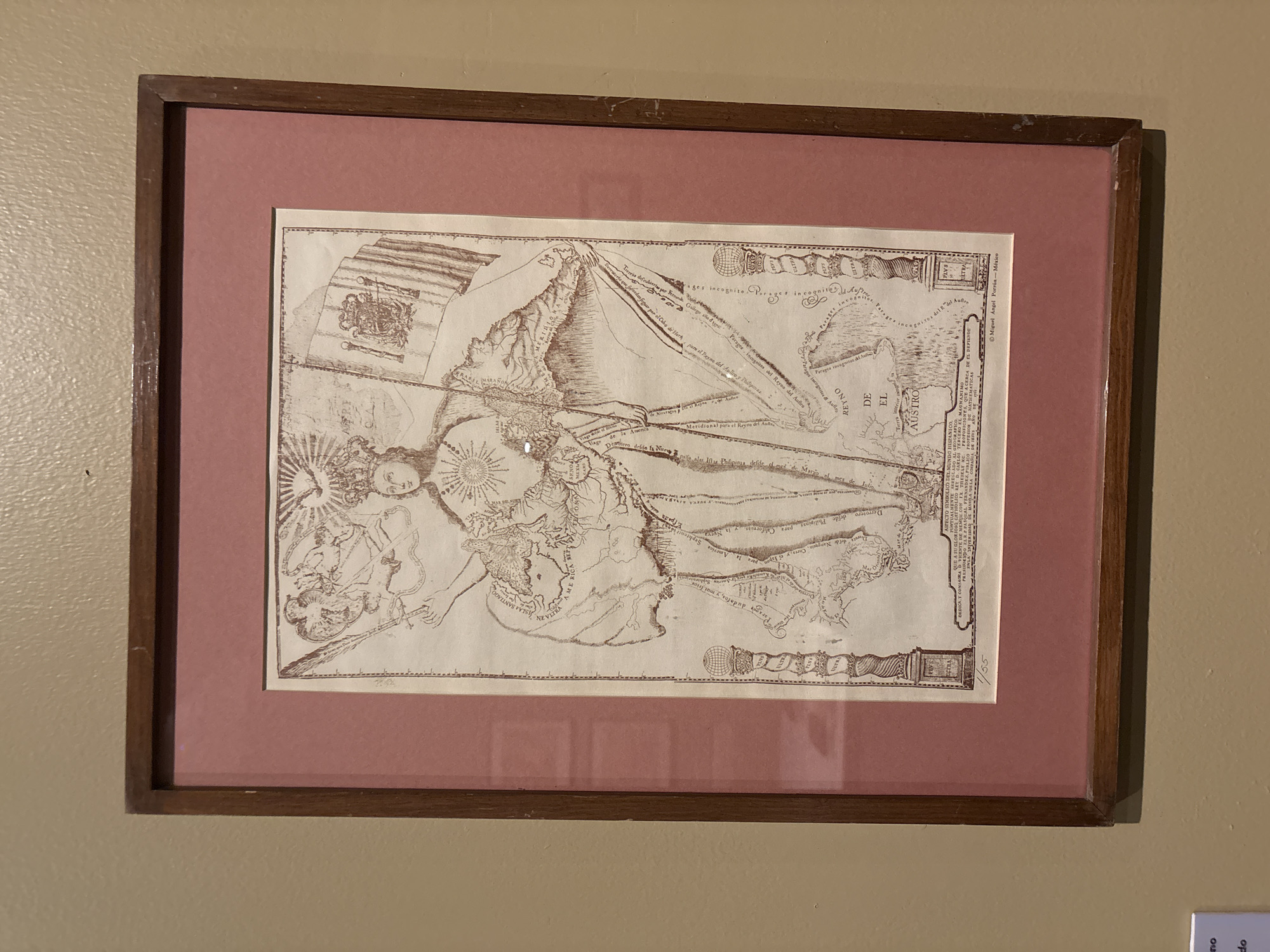 Here is a detailed description of the image:
The image shows a framed print on a light beige wall. The print depicts an antique map of the Americas, prominently featuring the western coastline of North and South America. The map is intricately detailed, with illustrations and text. The upper left corner includes a depiction of a person or figure. The frame is brown wood, and the print is mounted on a salmon-colored matte board. Small text in the lower right corner includes "Miguel Angel Porrúa- México". A fragment of a white label with the text "do" can be seen in the bottom right corner of the image.