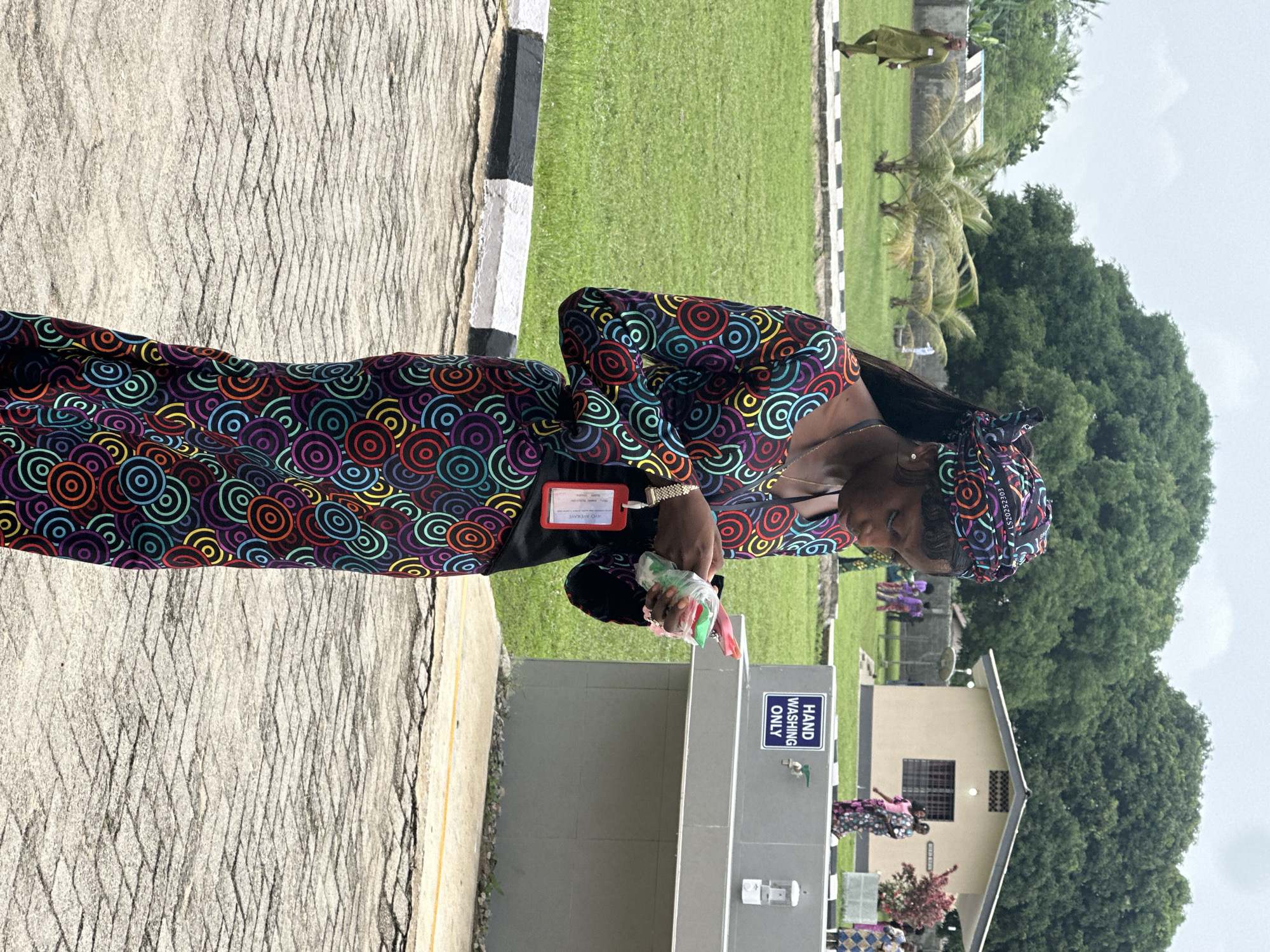 A young Black woman, wearing a colorful, circular-patterned dress and headscarf, stands on a paved surface and examines a small, clear plastic bag containing some items in her hands. She has a red identification badge attached to her dress. Behind her is a gray handwashing station with a sign that reads "HAND WASHING ONLY." Further in the background, several other people in traditional African attire are visible near a light-colored building. The setting appears to be an outdoor, possibly rural, area with green grass and trees. The sky is overcast.