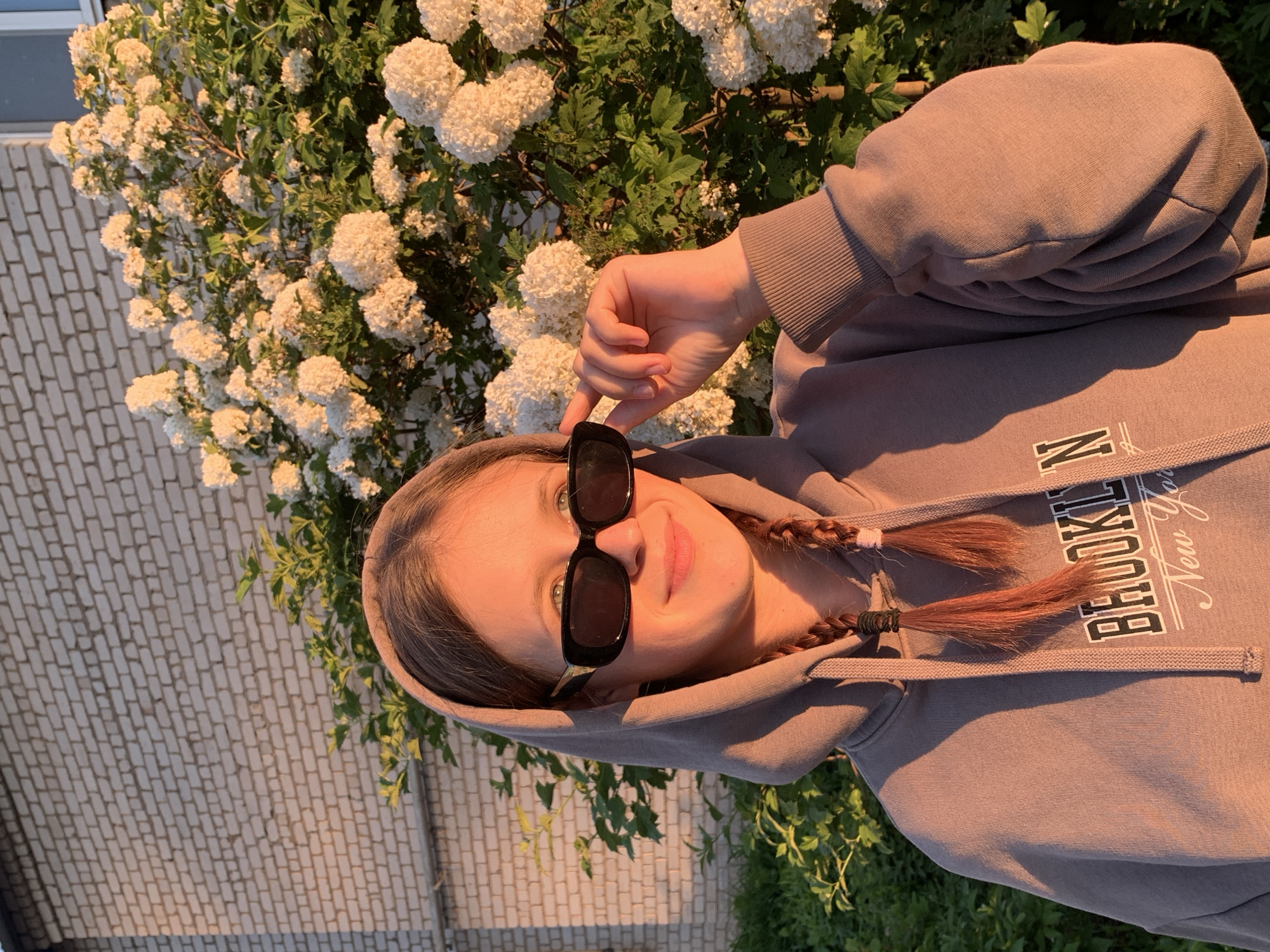 A young person, likely female, is shown in a vertical-oriented, eye-level shot, smiling and looking towards the camera while subtly adjusting their black rectangular sunglasses. They are wearing a greyish-brown hooded sweatshirt (hoodie) with the text "BROOKLYN New York" printed on the chest in white letters. Two small, reddish-brown braids are visible, emerging from the hoodie.
The individual is positioned outdoors, set against a backdrop of a large, lush bush or tree profusely covered in clusters of small, white flowers, suggesting springtime. To the left, a light-colored brick wall is visible. Green foliage can be seen beneath the flowering bush.
The scene is illuminated by warm, golden light, indicative of late afternoon or early evening during the "golden hour," creating soft highlights and gentle shadows. The weather appears sunny and clear. The person's expression conveys a sense of happiness or ease. The location is Фоки, Russia.