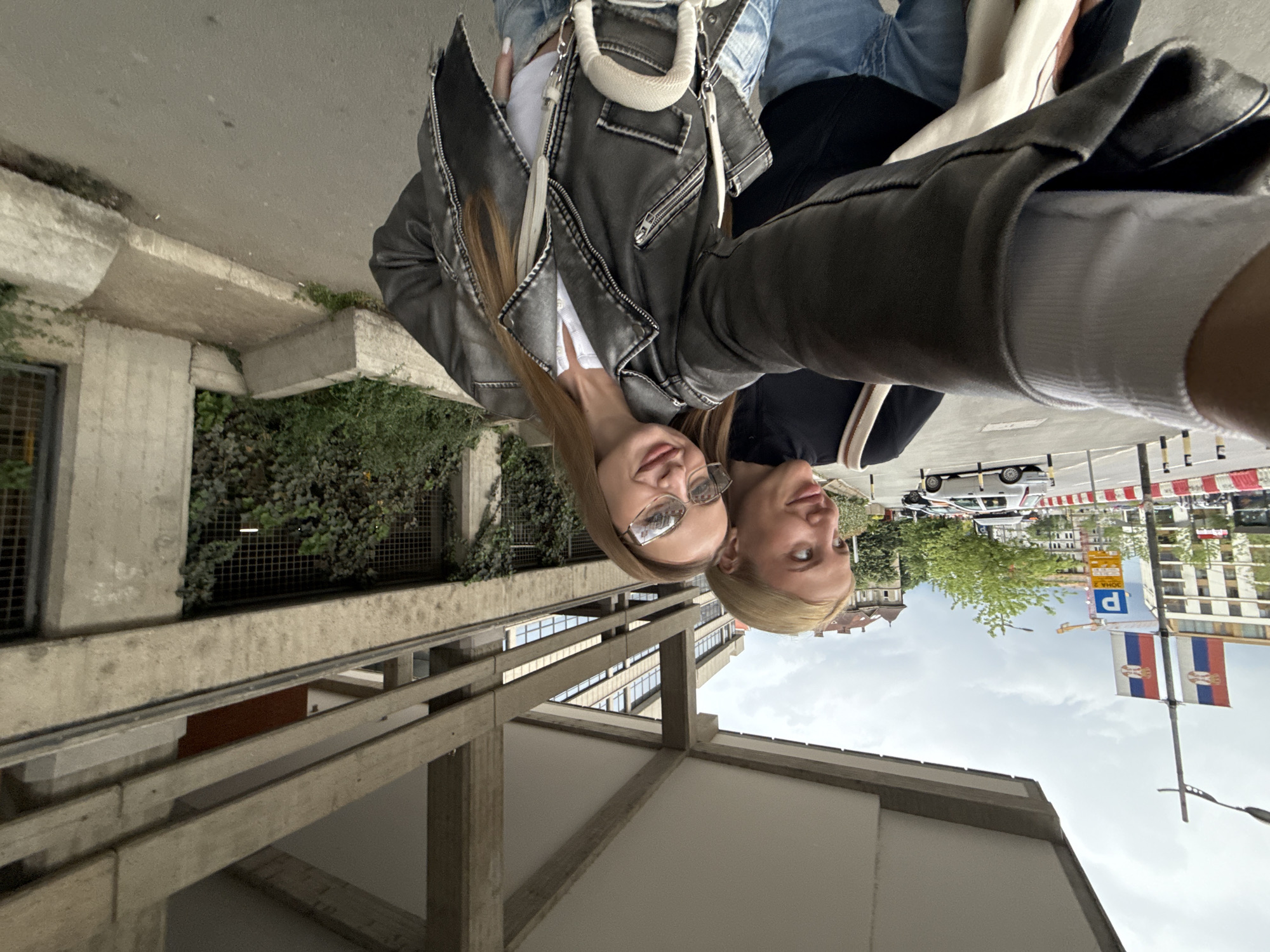 Two young women are posing for a selfie outdoors. One woman has long light brown hair and is wearing glasses, a white top, and a gray leather jacket with a white purse. The second woman has short blonde hair and is wearing a black top with white trim. They are standing in front of a concrete structure with green foliage and metal grates. In the background, a street with cars, buildings, and Serbian flags are visible. Based on the flags and architecture, the location is likely Belgrade, Serbia.