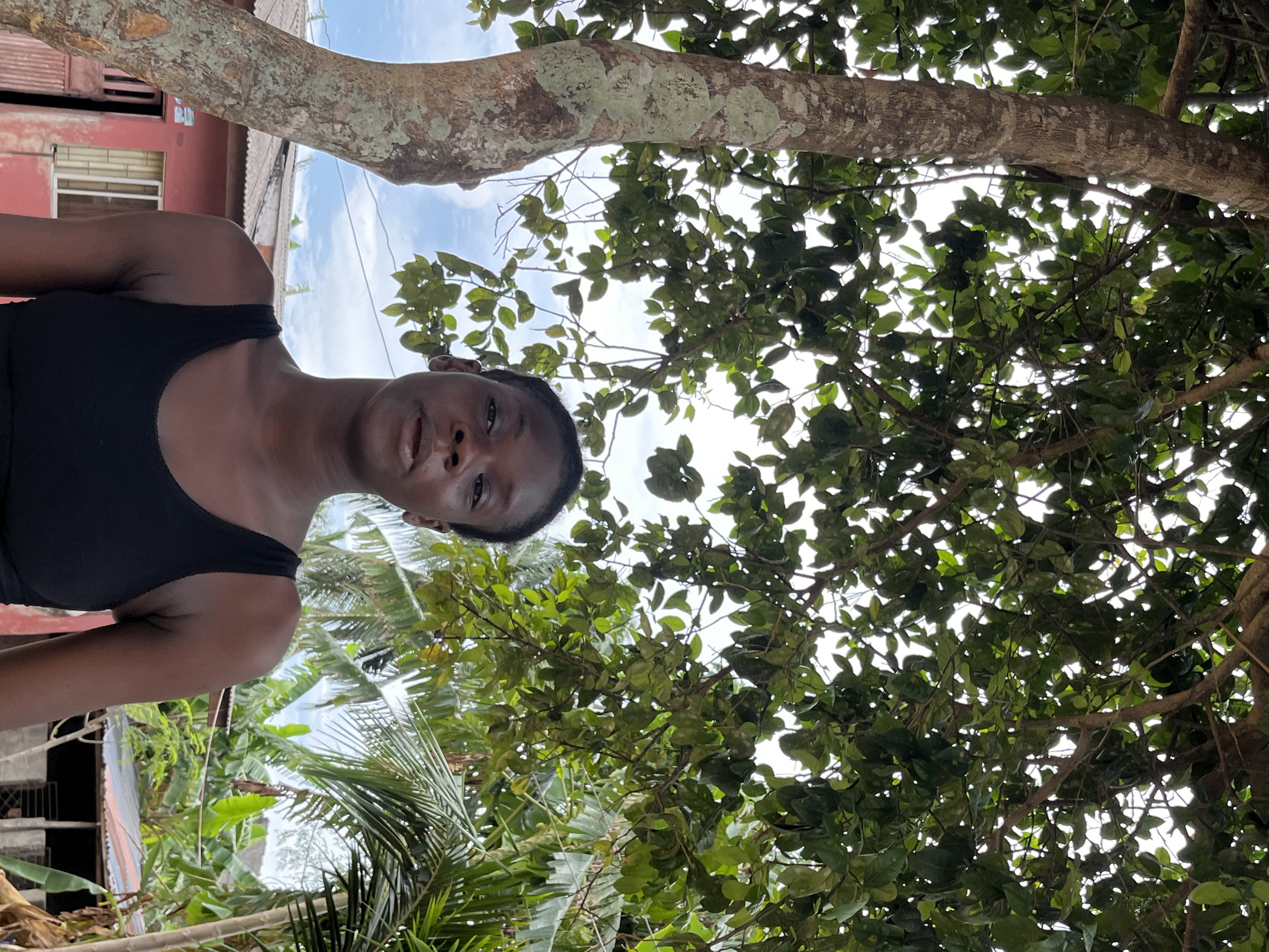 A photograph taken in Badagry, Nigeria, shows a Black woman wearing a black tank top. She is standing outside with lush greenery surrounding her. A thick tree branch with green foliage extends across the top of the frame. In the background, there is a building. The sky is visible through the branches of the tree. Palm trees and other tropical plants are also visible.