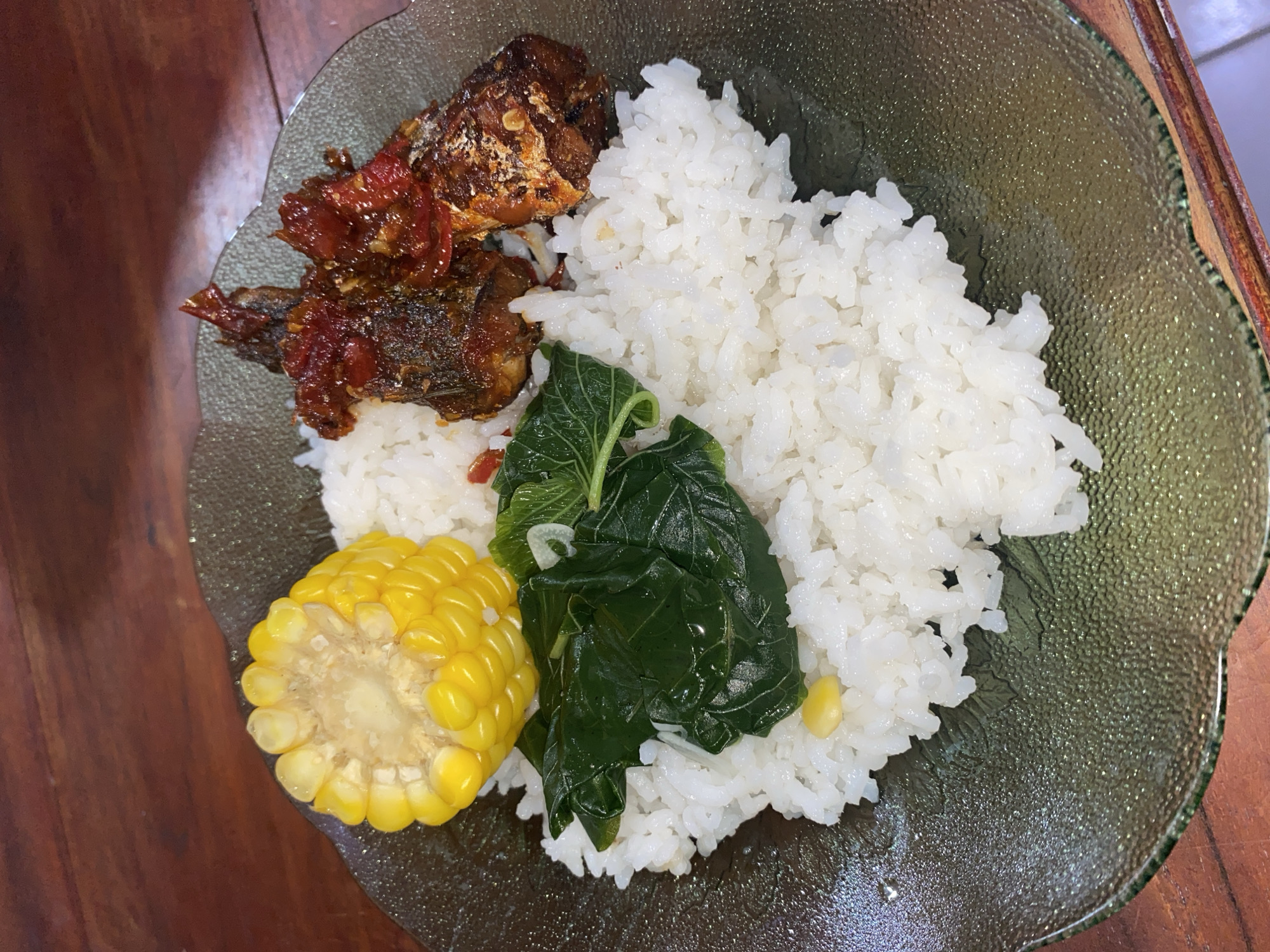 A close-up, overhead shot features a plate of food on a wooden table. The plate, which appears to be made of dark green, textured glass, contains a serving of white rice. Arranged on top of the rice are several items: a piece of cooked corn on the cob, a cluster of cooked green leafy vegetables, and several pieces of fried fish covered in a reddish sauce. The wooden table has a visible grain and is a warm brown color. The overall lighting is dim, suggesting an indoor setting. The image's composition places the food as the central focus, with no people or other identifiable objects in the frame. The location in Ta', Indonesia is not visually discernible from the image itself.
