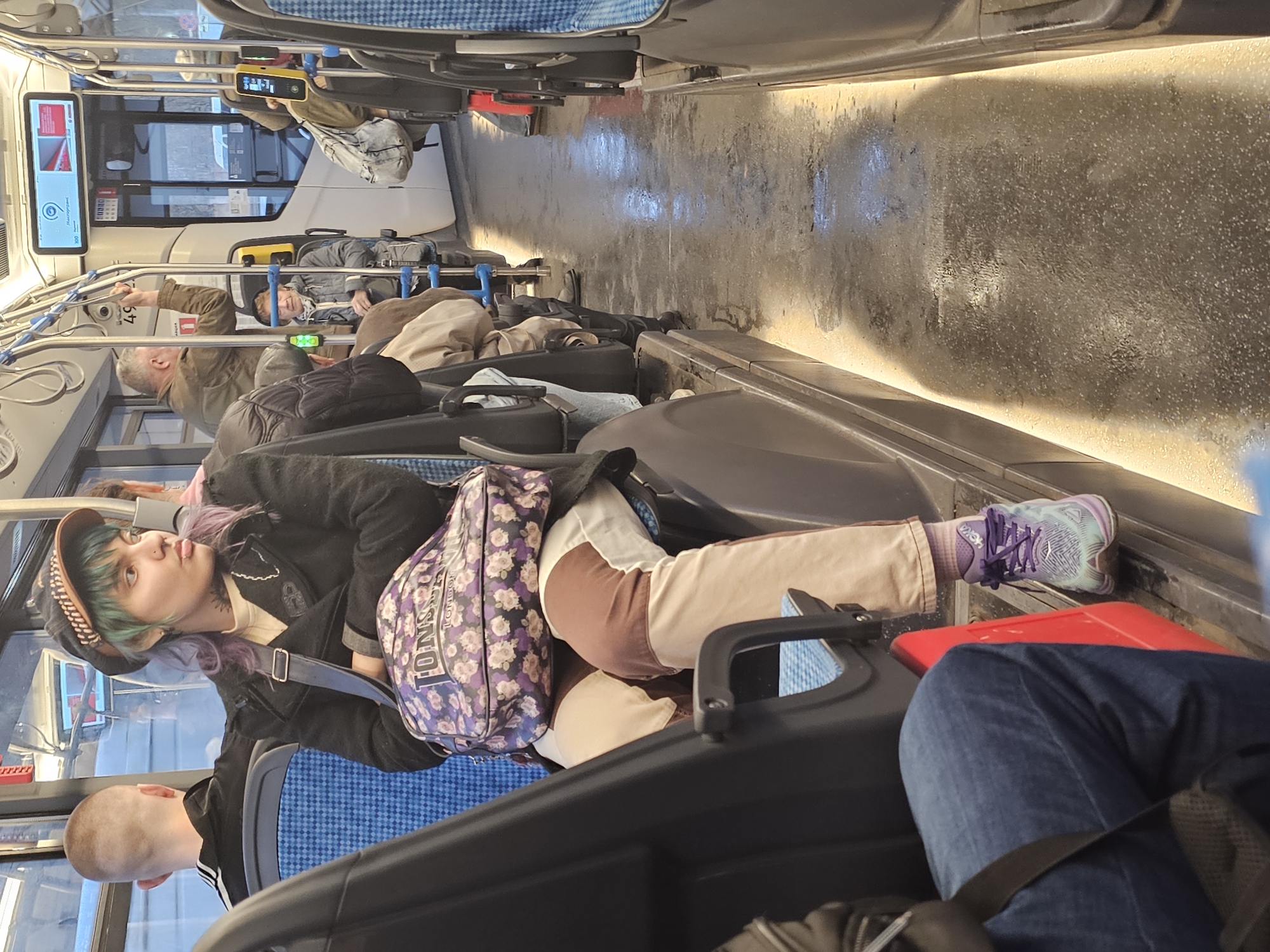 The photo appears to be taken inside a public transit vehicle, likely a bus or tram, in a city. Passengers occupy the seating, with several people visible. One individual with dyed hair, a cap, and a shoulder bag is prominently featured. Other people are seated nearby and further in the background of the vehicle. The interior environment shows the typical layout of seats, windows, and overhead structures in a public transit setting. The city of Khimki, Russia is not deducible from the image.