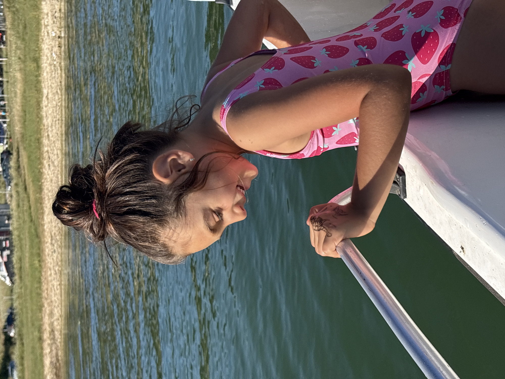 A young girl with dark hair pulled back in a bun is on a boat. She is wearing a pink swimsuit with a strawberry pattern. The boat is on the water, and a sandy shore with green vegetation is visible in the background. The sun is shining, casting a shadow on the girl's legs. The girl appears happy, with a smile on her face. The water is a deep blue-green. The scene suggests a sunny day, likely in the warmer months, possibly a vacation or day trip.