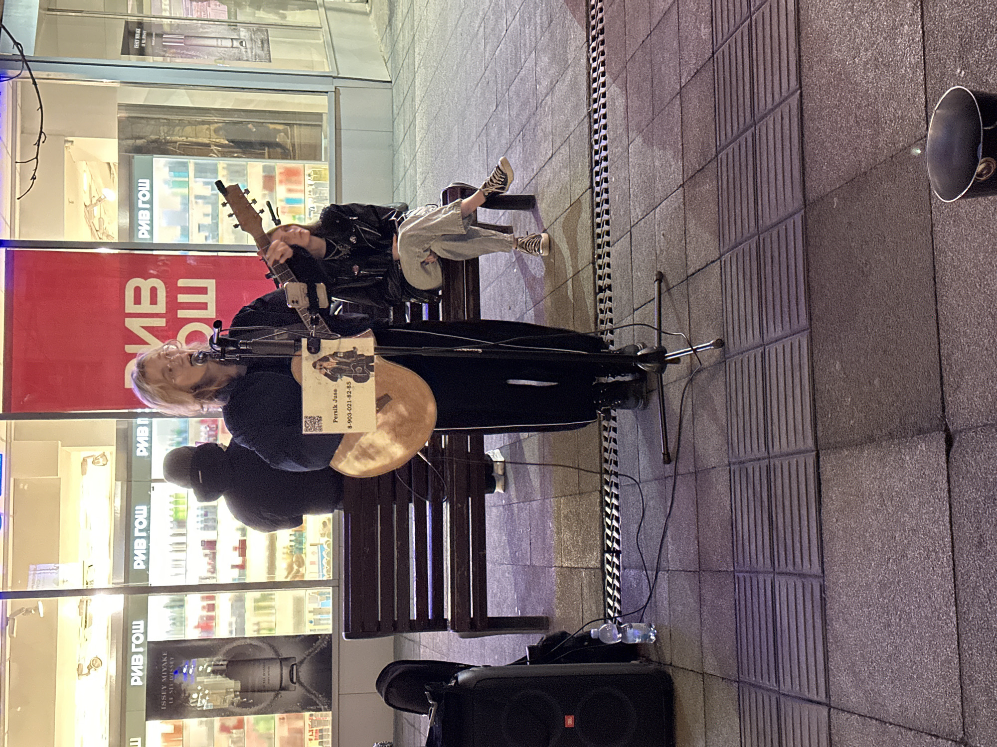 On a street in Saratov, Russia, a street performer stands on a paved surface next to a park bench, a PA speaker, and a cymbal. The performer sings into a microphone and plays a stringed instrument. A sign with the name "Persik Juss" and a phone number sits on the microphone stand. Another person is sitting on the bench to the upper-right of the performer. In the background is a storefront for a beauty store named "РИВ ГОШ", with a display of "ISSEY MIYAKE" products. A security mirror is fixed to the wall on the right.
