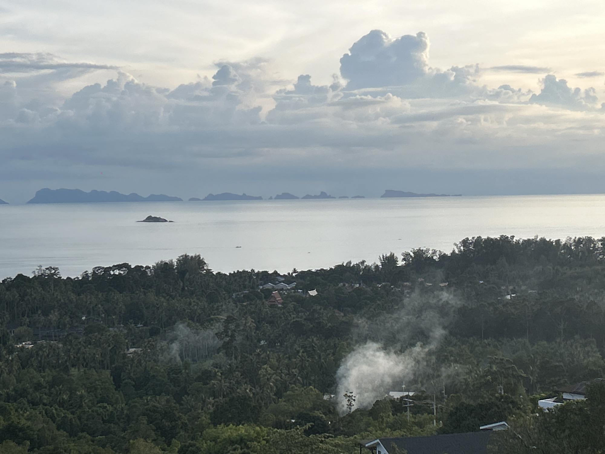 This image depicts a wide, panoramic view of a coastal landscape on Koh Phangan island in Thailand. The scene is divided into three main horizontal bands: the sky, the sea, and the land.
The sky is overcast with a soft, diffused light, suggesting either early morning or late afternoon. Large, billowing clouds dominate the upper portion of the image. Some are dark and dense, while others are lighter and more translucent, allowing a pale yellow light to filter through from the horizon.
The sea occupies the middle band. It appears calm and vast, stretching towards a distant horizon. A chain of islands or peninsulas forms a faint, silhouetted outline against the horizon. One small, isolated island with a distinct peak stands out in the mid-distance on the left side of the frame. A few small boats are visible as tiny dots on the water.
The foreground is a dense, verdant jungle covering rolling hills that slope down towards the sea. The foliage is predominantly lush green, with varying shades indicating different types of trees and vegetation. Scattered throughout the jungle are rooftops of houses and buildings, suggesting a sparsely populated area. A notable feature is the presence of white smoke or mist rising from within the jungle, possibly from a controlled burn or cooking fires. The smoke billows upwards and disperses among the trees, creating a hazy effect. The overall impression is one of a tropical, natural environment with a human presence integrated into the landscape.
