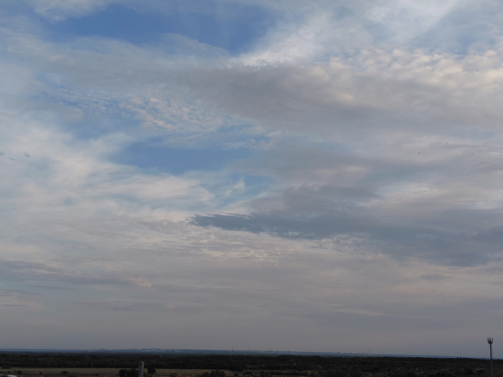 A wide, low-angle shot depicts a hazy sky filled with scattered clouds of varying densities, ranging from wispy cirrus to thicker, more opaque formations. The sky transitions from a pale blue near the top to a grayish-white hue closer to the horizon. Below the sky, a dark, dense band of trees stretches across the frame, representing a forested area. In the far distance, a faint, hazy line of what appears to be industrial structures or a city skyline is visible along the horizon. On the far right side of the image, a tall, slender cellular tower with antennas stands out against the sky and trees. The overall impression is a natural landscape with a distant urban or industrial element under a cloudy sky. The location is the city of Samara, Russia.