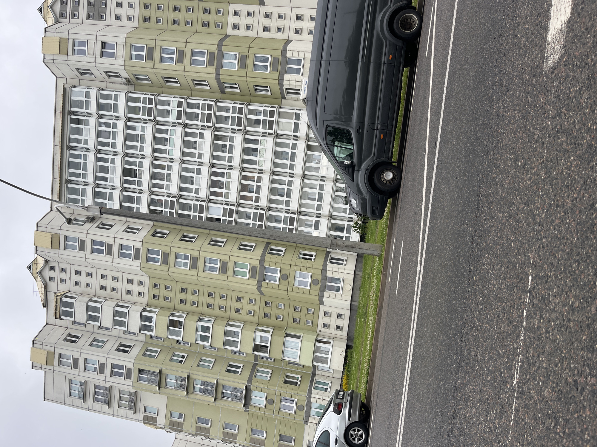 The image depicts a street view in Minsk, Belarus, featuring a large, modern apartment building with a distinctive wavy facade. The building's exterior is a combination of light green and beige panels, with numerous white-framed windows, some of which are balconies. To the right of the apartment building, another building with a more traditional gray facade and white windows is visible. In the foreground, a dark grey van is parked on the side of an asphalt road, which has white lane markings. A silver car is parked further down the street to the left of the van. The sky is overcast, suggesting a cloudy day. The scene appears to be a residential area in the city.