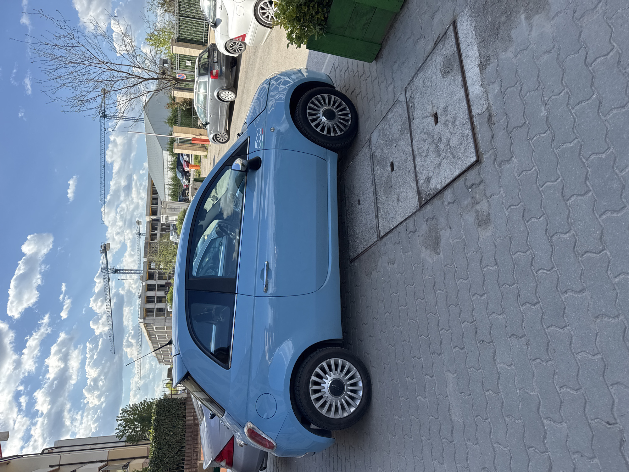 The image depicts a light blue Fiat 500 parked on a paved street with interlocking pavers in Sofia, Bulgaria. The car is situated next to a square concrete manhole cover framed with metal. In the background, there are construction cranes visible against a partly cloudy sky. Other parked cars, including a grey wagon, are also present. The environment suggests an urban setting.