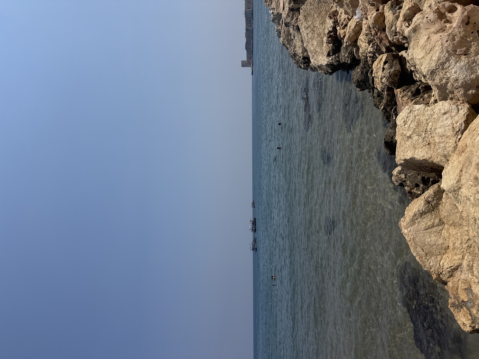This is a wide shot of the Mediterranean Sea in Erdemli, Turkey. In the distance, a fortified island structure, likely a castle or a historical monument, stands on a small peninsula. Several boats are visible on the water. In the foreground, large rocks line the shore, with some areas of clear, shallow water where the seabed is visible. A few people can be seen swimming or wading in the sea. The sky is clear and blue.