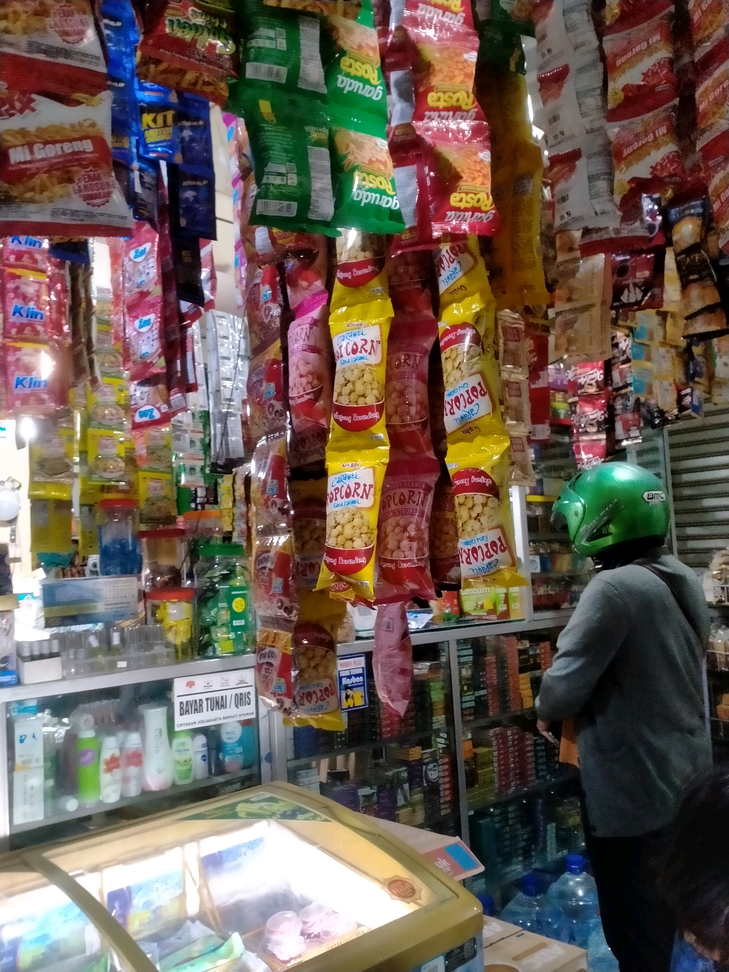The image depicts the interior of a small convenience store located in Kasihan, Indonesia. The store is densely packed with merchandise, primarily snacks and toiletries, displayed on hanging racks and shelves. Products include various packaged snacks, such as "Mi Goreng" and popcorn, as well as bottled liquids and consumer goods. A person wearing a grey jacket and a green helmet is standing at the counter, facing away from the camera, with their back to a display of goods. Another person's head is visible in the lower right corner of the frame. A glass-fronted refrigerator or freezer is visible in the foreground, containing packaged food items. The lighting is dim, with some overhead illumination contributing to the overall ambiance of a busy local shop.
