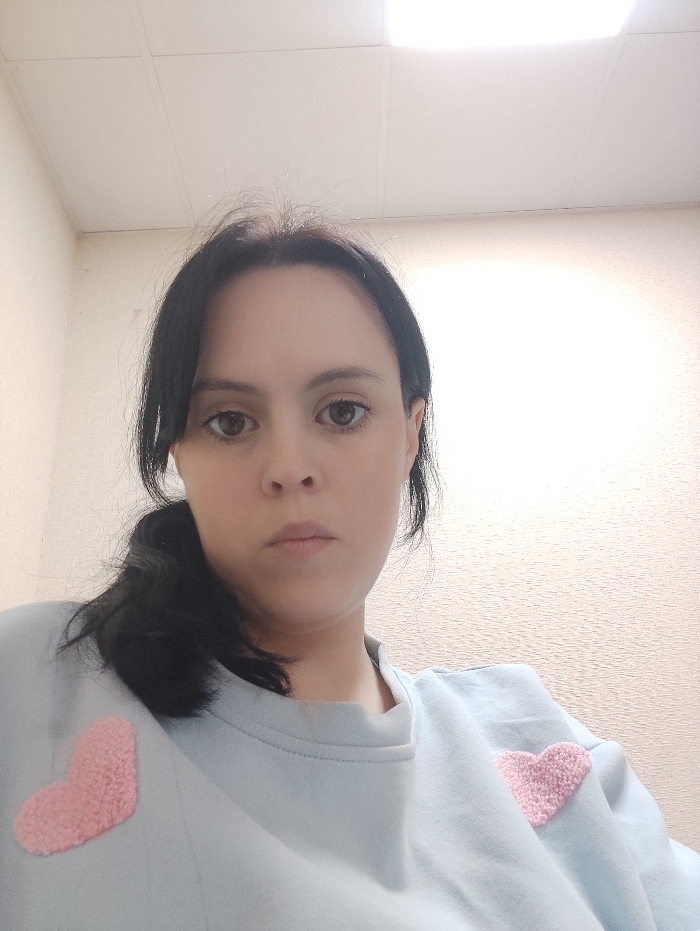 The image displays a female individual, appearing to be an adult, positioned centrally in an indoor environment. She possesses dark hair, styled into a ponytail on her left side, and brown eyes directly engaging the camera with a neutral facial expression. Her attire consists of a light blue long-sleeved sweatshirt, featuring two distinct pink, textured, heart-shaped appliques, one on each side of the chest. The background reveals light-colored, subtly textured walls and a white suspended ceiling with visible grid lines. Two recessed rectangular light fixtures are present in the ceiling, emitting bright overhead illumination, with one fixture on the right exhibiting a slight glare. The photograph is captured from a slightly upward angle. No specific visual information is present within the image to determine the exact location within the city of Donetsk, Ukraine.