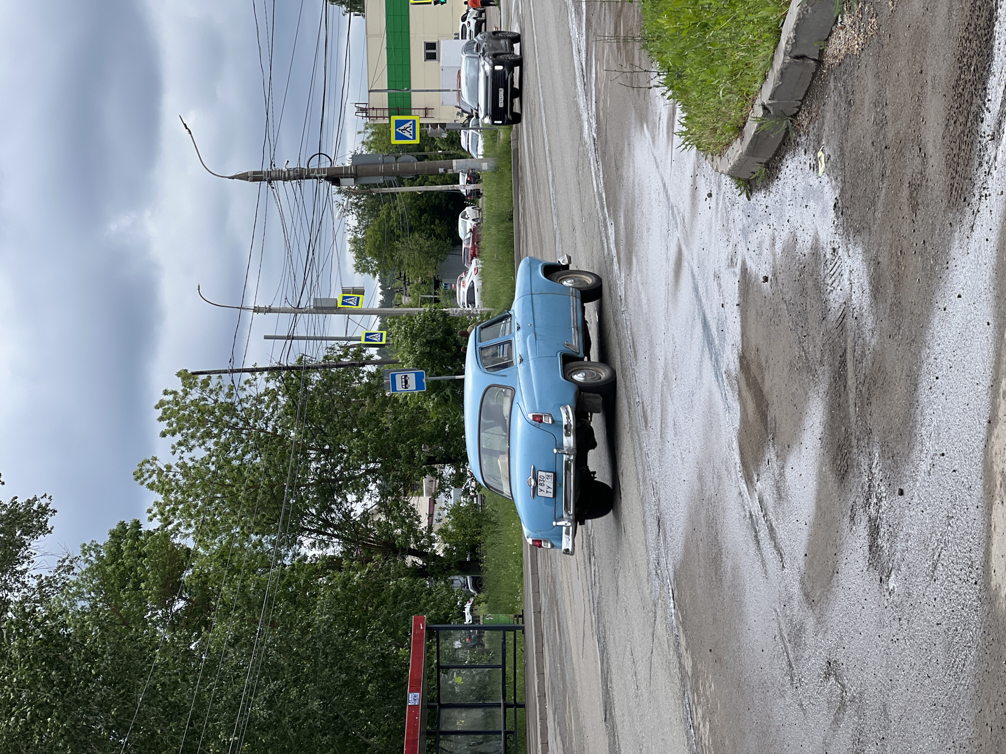 The image depicts an urban street scene in Izhevsk, Udmurt Republic, Russia, identified by the "18" regional code on the visible vehicle license plates and the characteristic infrastructure. The scene is captured on an overcast day, with wet asphalt and visible puddles suggesting recent rainfall.
The primary subject is a light blue vintage sedan, consistent with a GAZ-21 Volga model, driving away from the viewer in the left lane. Its rear features a chrome bumper and a Russian license plate displaying "У 830 ТУ 18". On the left side of the road, a bus stop shelter with a red frame and glass panels is present, with a blue rectangular bus stop sign mounted on an adjacent pole. A person is partially visible within the bus stop shelter.
In the midground, a dark-colored SUV, bearing Russian license plate "С 674 УУ 18", is approaching the viewer in the right lane, on the opposite side of the street from the blue sedan. An intersection is visible further down the road, with additional vehicles parked and in motion.
The street is lined with numerous utility poles supporting a dense network of overhead electrical and communication wires. At least three yellow triangular signs indicating pedestrian crossings are affixed to these poles. Streetlights are also mounted on some poles. The road surface in the foreground and midground is wet, dark asphalt with visible cracks and areas of standing water. A concrete curb delineates the right edge of the road, alongside which a strip of green grass and weeds grows.
The background is dominated by a dense canopy of green deciduous trees. Behind these trees and the utility infrastructure, several buildings are visible, including one with a light-colored facade featuring a horizontal green strip, and another with an external ladder visible on its side. The sky is uniformly grey and overcast.