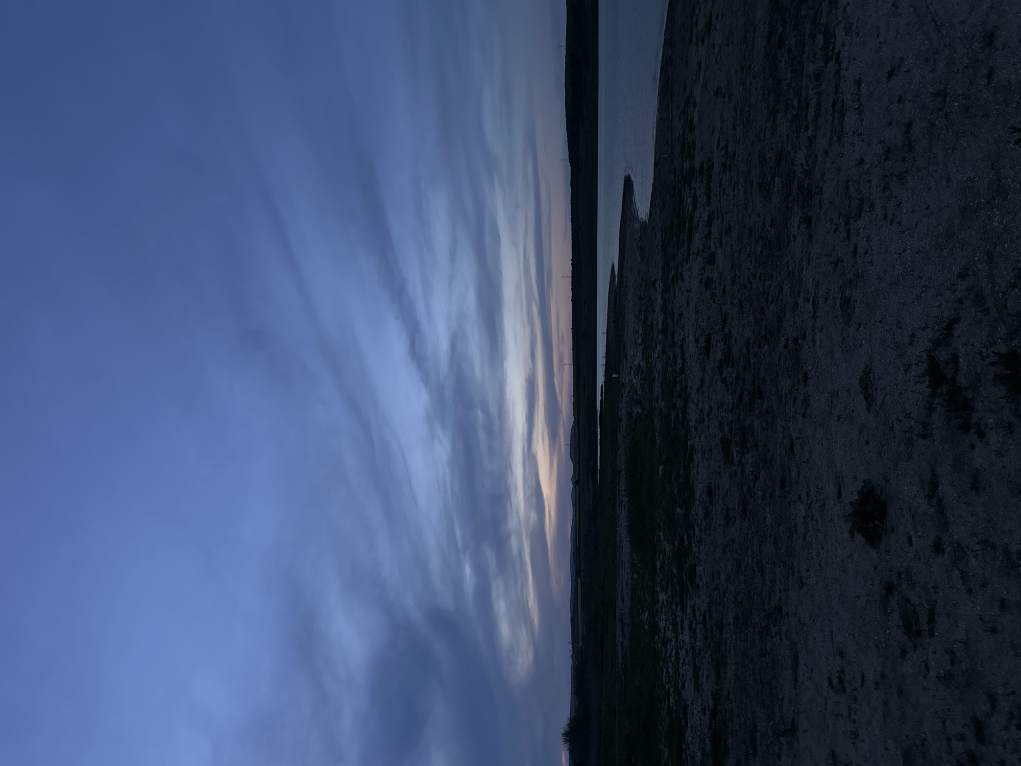 A vertical, long shot shows a twilight scene, likely in Trastikovo, Bulgaria, showcasing an overcast sky in gradations of blue and gray. The sky transitions to a slight orange tint along the horizon where it meets a body of water to the right. Adjacent to the water is a dark, indistinct shoreline or bank, which could be vegetation or rocky terrain. Details of the land are obscured due to the low light conditions. No people, specific objects, actions, or interactions are visible. The environment dominates the frame, depicting a natural setting at dusk or dawn.