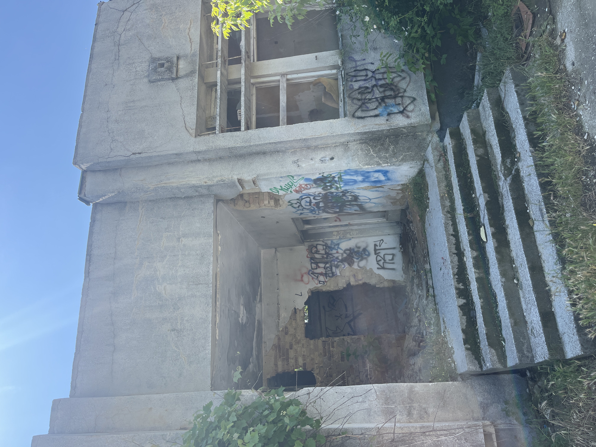 A decayed two-story concrete building stands on a bright, sunny day. Graffiti covers parts of the exterior walls, especially around the window and doorway openings. The structure is in a state of disrepair, with broken windows and damaged walls. A set of concrete steps leads up to the building. Plants and weeds grow around the building and stairs.
It is not possible to determine the location of the building is in Varna, Bulgaria from this image.