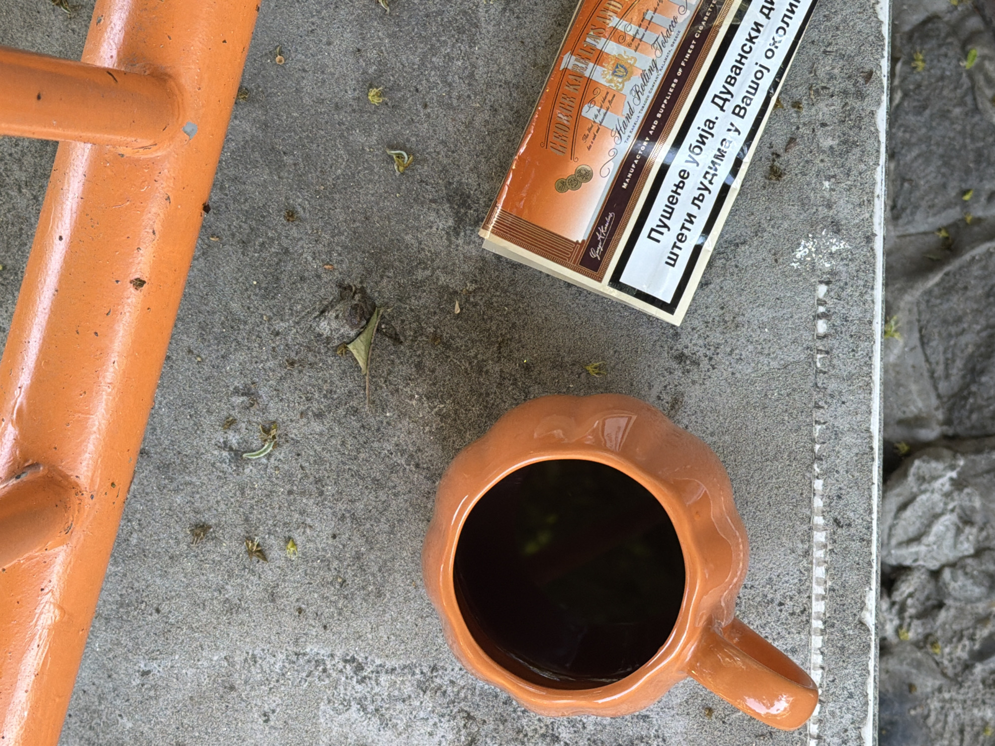 The image shows an outdoor scene on a concrete surface. An orange metal structure is on the left. A package of tobacco with text in Greek and Serbian lies near the top center. An orange mug filled with a dark liquid is near the bottom. The location is likely in Belgrade, Serbia, due to the Cyrillic script on the tobacco packaging. No people are visible.