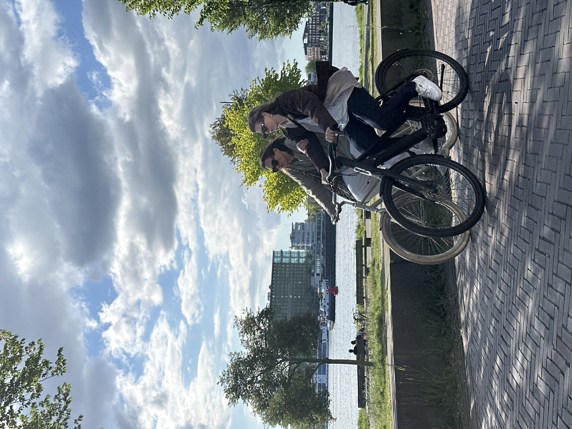 Two women are riding bicycles on a paved path alongside a body of water. Both women are wearing sunglasses. One is wearing a brown jacket and blue jeans, while the other is wearing a brown jacket and khaki pants. In the background, buildings can be seen across the water. Trees are visible along the path and in the background. The sky is cloudy. This scene is likely taking place in Amsterdam, Netherlands, due to the presence of canals and bicycles, which are commonly associated with the city.
