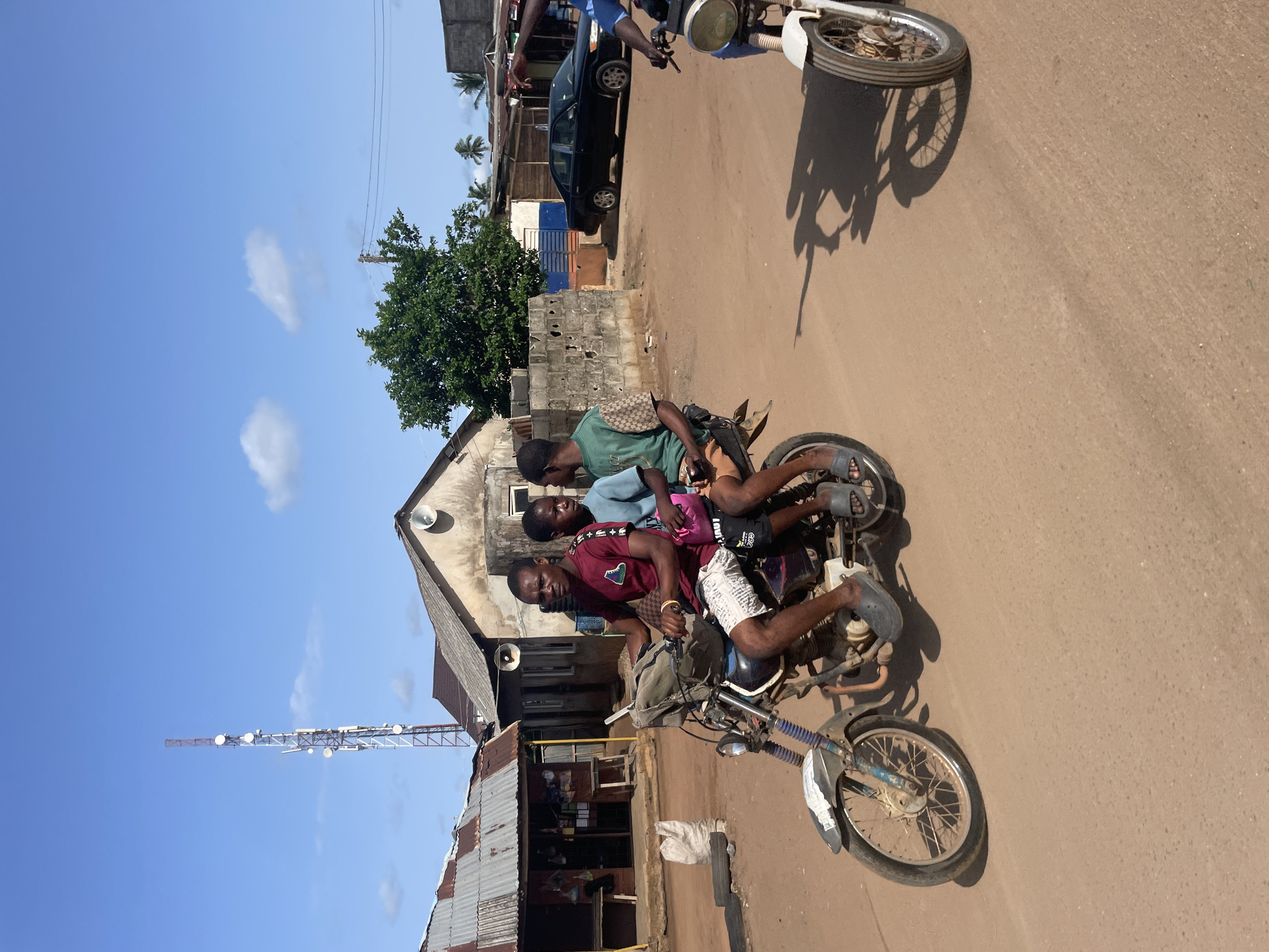 Three people are riding on a motorcycle on a dirt road. They are near a building with a corrugated metal roof and a communications tower. A car is parked along the road. The location appears to be in Ipota, Nigeria.