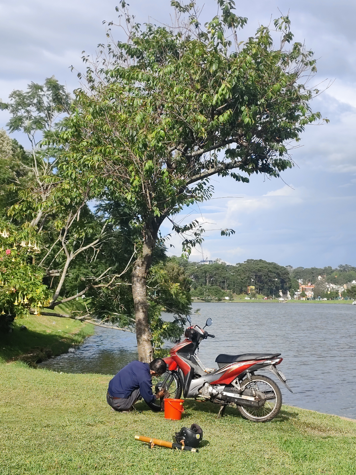In Da Lat, Vietnam, a man in a dark blue shirt and grey pants kneels on a grassy bank beside a tranquil lake. He is engaged in cleaning the wheel of a red and silver motorcycle parked nearby. An orange bucket sits beside him, and a black helmet rests on the grass in front of the motorcycle. The scene is set under a cloudy sky with a backdrop of lush green trees and distant buildings. The natural light suggests it is daytime.