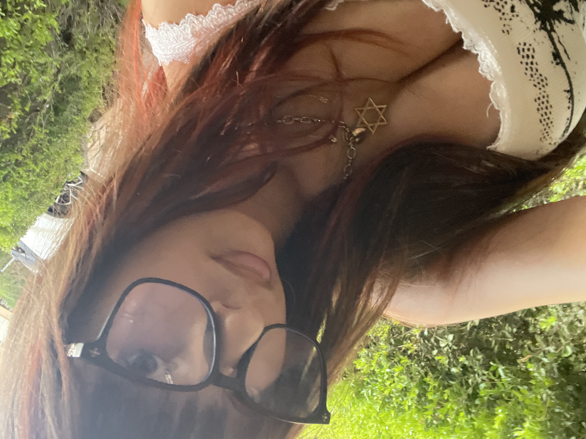 A young woman with long, dark brown hair with reddish highlights is taking a selfie outdoors. She is wearing large, black-rimmed glasses and a white top with black patterns and lace trim. A silver necklace with a Star of David pendant is visible. The background consists of lush green foliage and a blurred suggestion of a car in the distance. The lighting suggests it is daytime. The woman's face is partially obscured by her hair and glasses, and she is looking towards the camera with a slight smile.