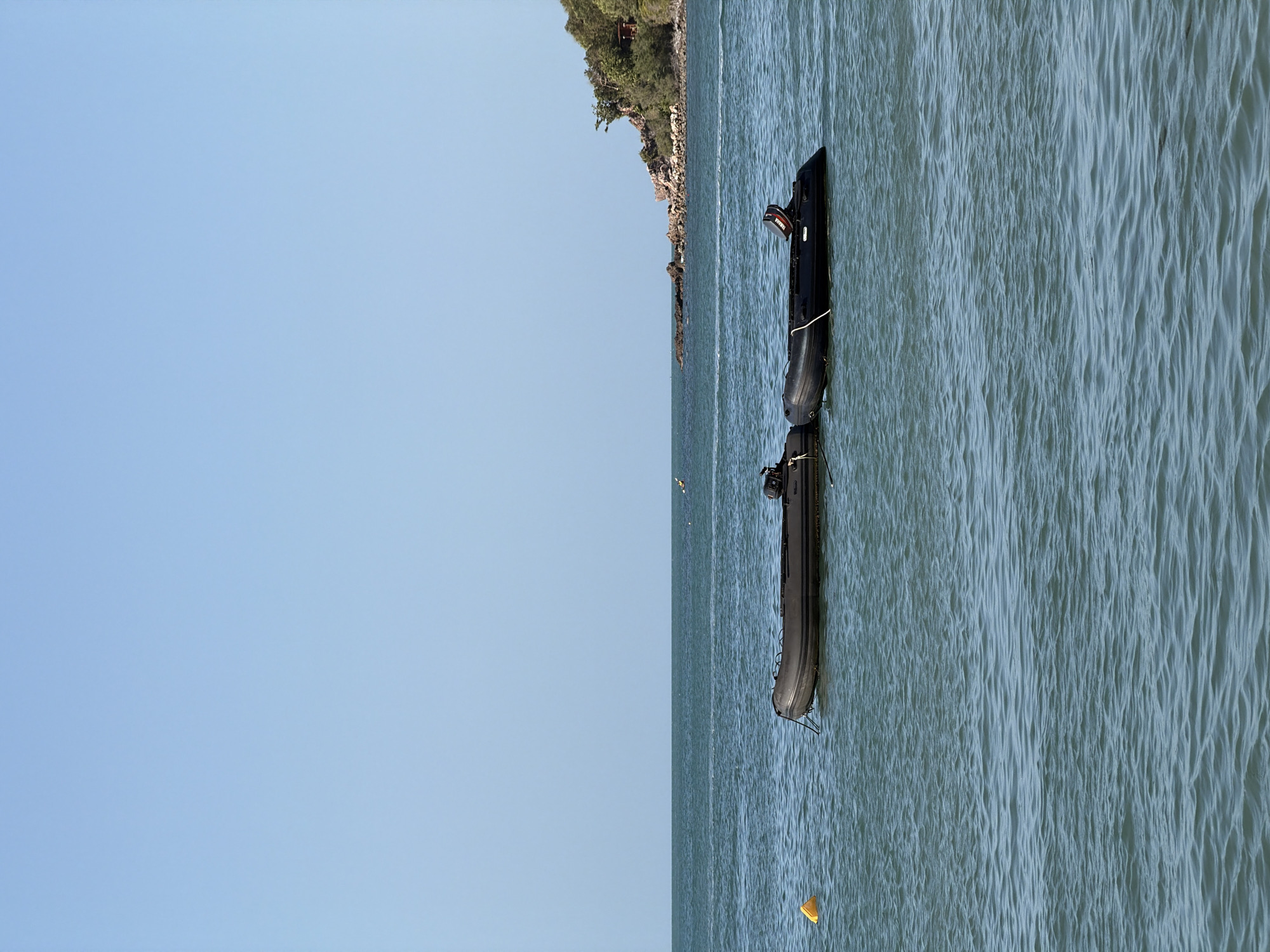 Two inflatable boats are moored in the ocean, with a clear blue sky overhead and a distant, tree-covered island on the horizon. One boat is dark gray, and the other is black with a Yamaha outboard motor visible at the stern. A yellow buoy floats in the water to the left of the boats. The scene appears to be a calm day in a coastal area of Ko Samui District, Thailand, with the gentle waves indicating a tranquil marine environment.