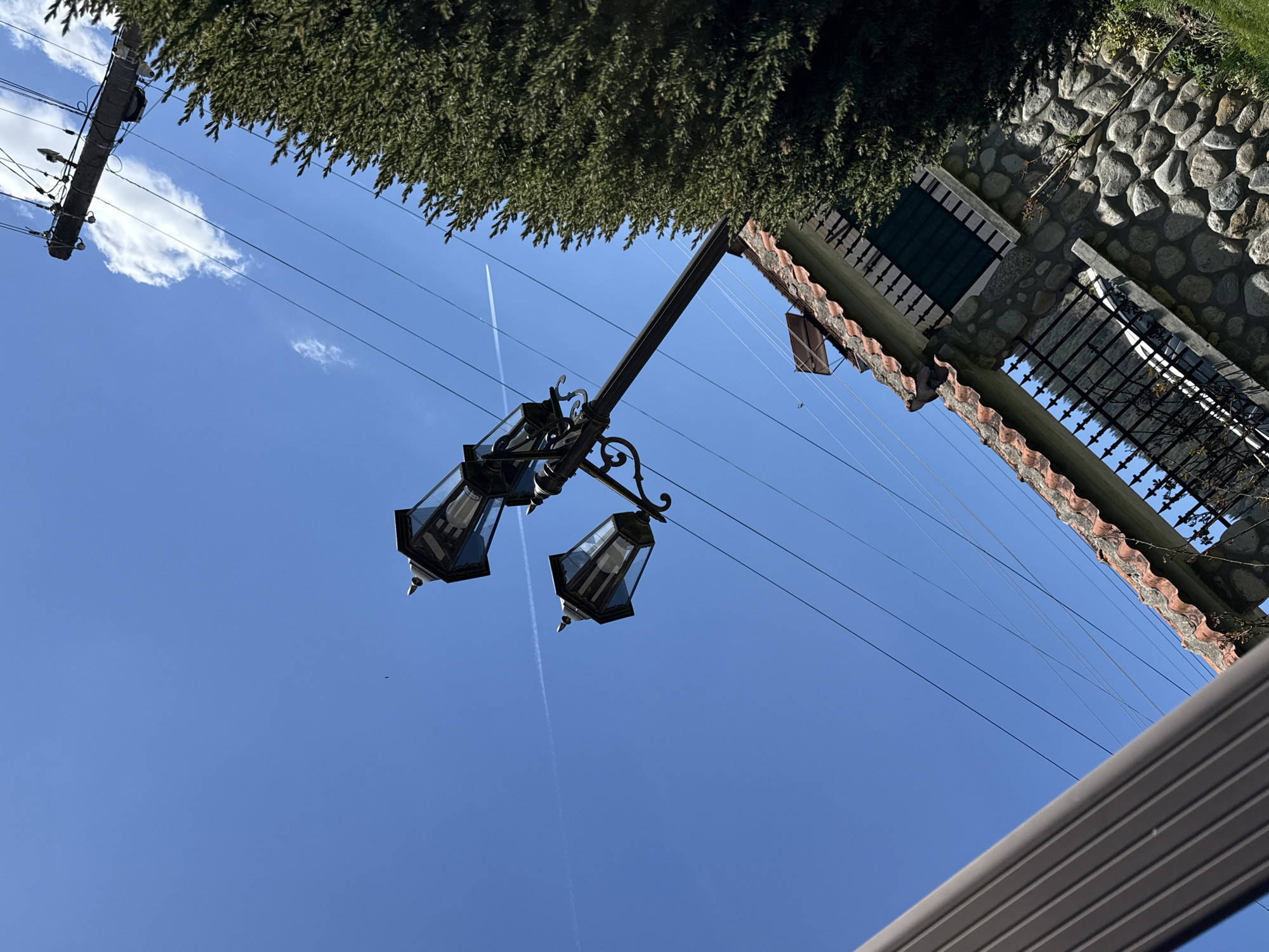 The image, taken from a low angle, features a clear, blue sky above Beli Iskar, Bulgaria. A contrail bisects the sky. A vintage-style lamppost with two lanterns is in the foreground. Overhead power lines run across the frame. A portion of a building with a stone wall and a red-tiled roof is visible on the right. Green foliage appears at the top of the image. A section of a gray, corrugated surface is at the bottom of the frame.