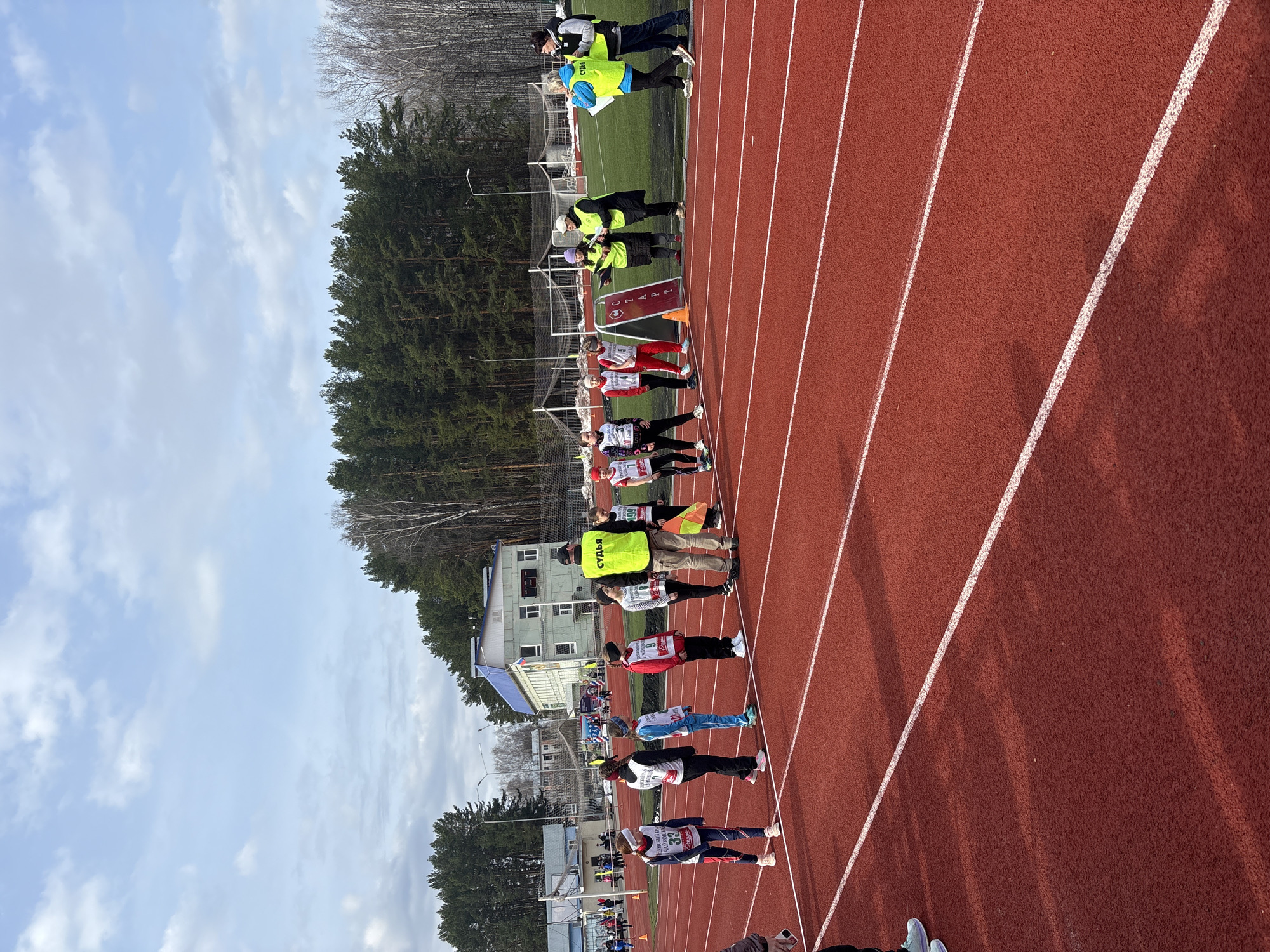 The image depicts a track and field competition, likely a race, taking place in Chaikovsky, Russia. Athletes are lined up at the starting line on a red track with white lane markers. Officials wearing neon yellow vests oversee the event. The background includes trees, a blue-roofed building, and a green field. The sky is partly cloudy. Competitors are wearing numbers on their chests.