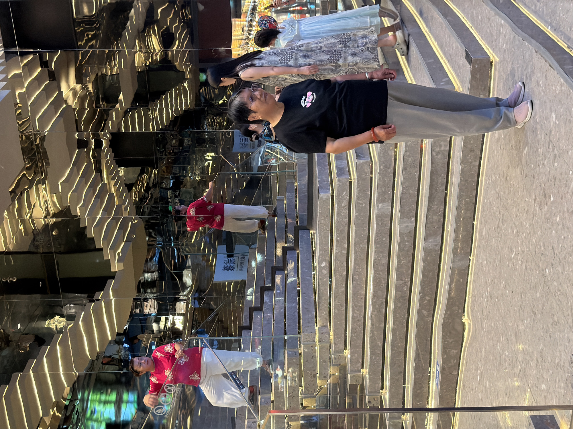 In Yuzhong District, China, a mirrored staircase is the backdrop for several individuals. A woman in a pink top and white pants stands on the second step, holding a rope. Further up, another person in a red top and white pants is seen on the stairs. In the foreground, a woman wearing a black t-shirt and gray pants with pink sneakers looks to the right. Behind her, a woman in a patterned dress and another in a light blue dress are ascending the stairs, with the latter holding a decorative object. The reflective surfaces of the staircase create a disorienting and layered visual effect.