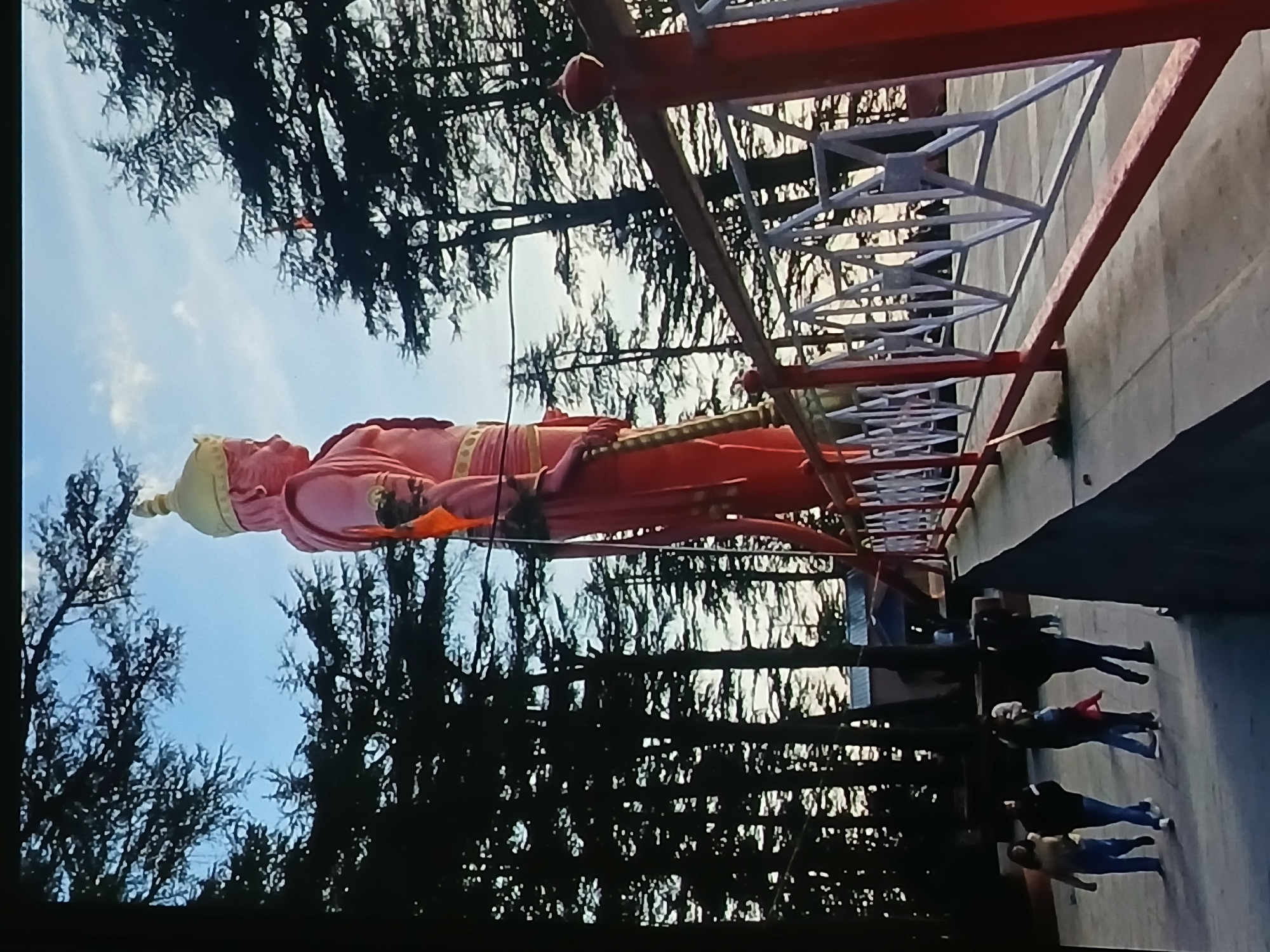 The image shows a large, pink statue of Hanuman in Bengaluru, India. The statue is surrounded by trees and a structure of red and white metal beams. Several people are walking on a pathway near the structure. The sky is visible in the background.