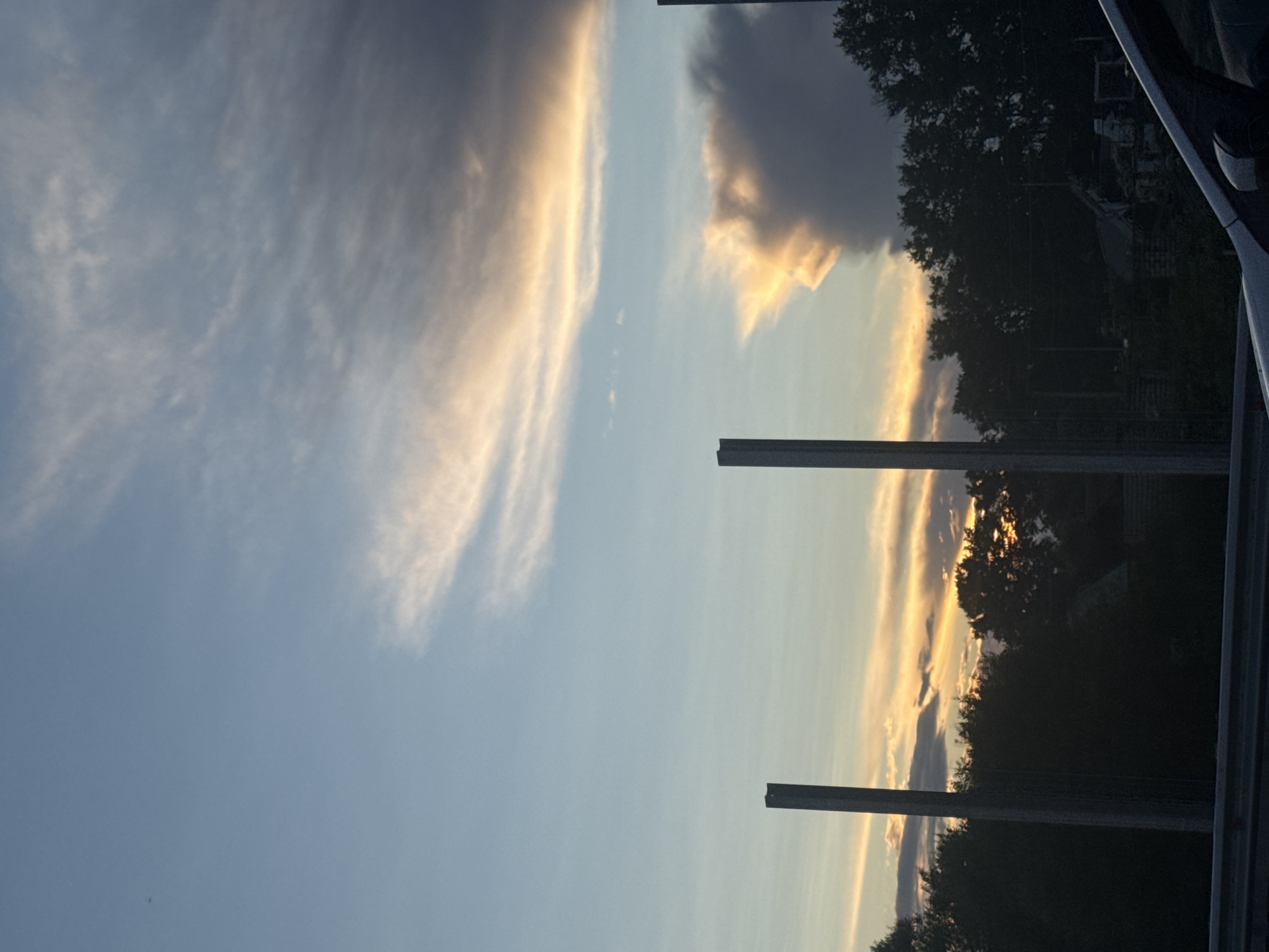 The image captures a twilight sky over Krutitsy, Russia, likely from the perspective of a vehicle. The sky is a canvas of soft blues and oranges, with wispy clouds streaked with sunlight, hinting at the setting sun. Two tall, dark, rectangular pillars, possibly part of a fence or structure, stand in the foreground, bisecting the view. Beyond them, a dense line of dark trees silhouettes the horizon. The overall atmosphere is serene, with the natural beauty of the sky contrasting with the man-made structures. There are no people visible in the frame.