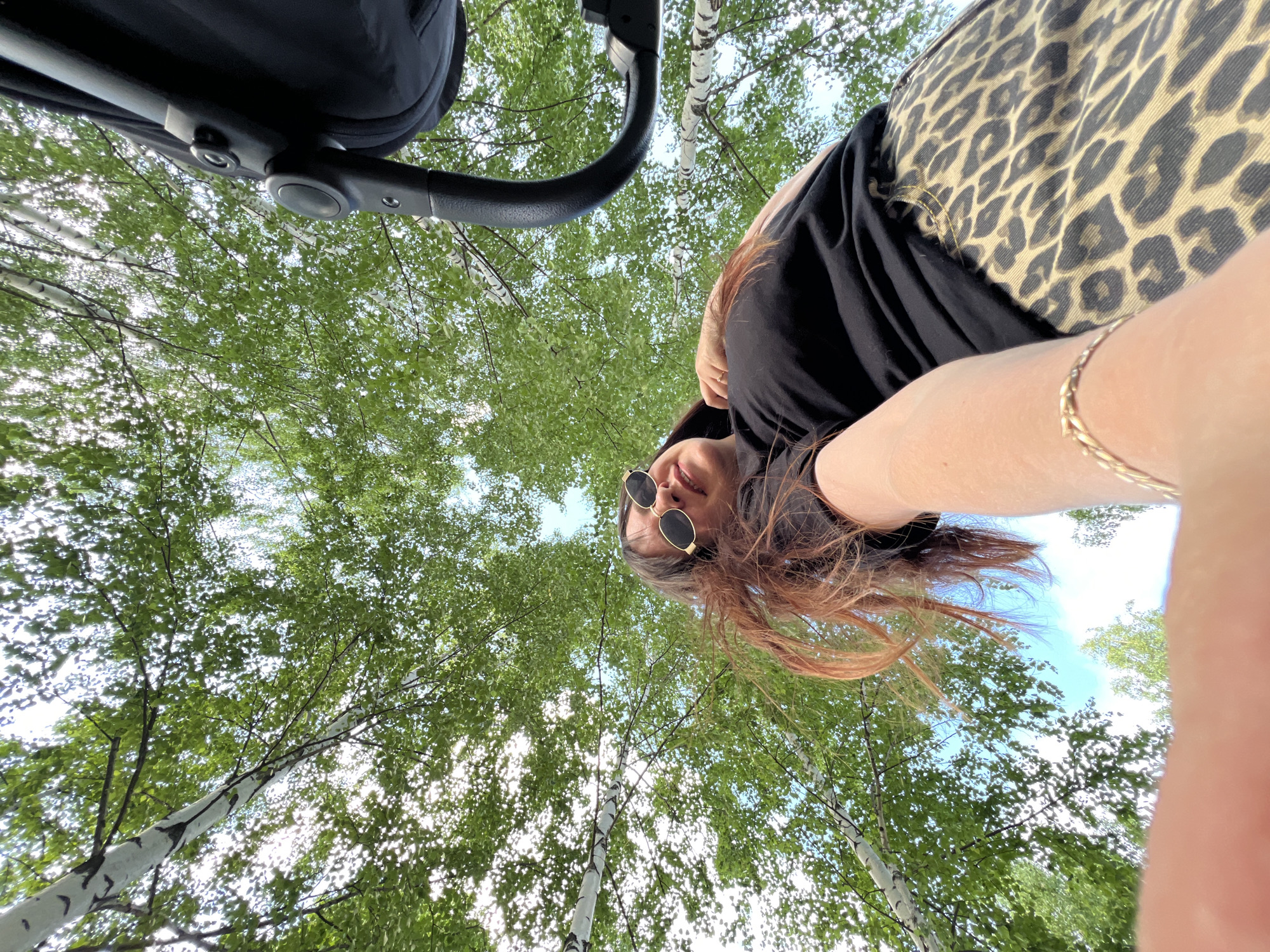 An outdoor, ground-level shot features a woman looking up at the camera from a low-angle perspective. She is wearing sunglasses and a black t-shirt paired with a leopard-print skirt. Her auburn hair is wind-swept, and a baby stroller is partially visible to the right. The background is dominated by the canopy of birch trees, with sunlight filtering through the green leaves, and a clear blue sky peeking through. The image appears to have been taken in a park or wooded area within Noviy, Russia.