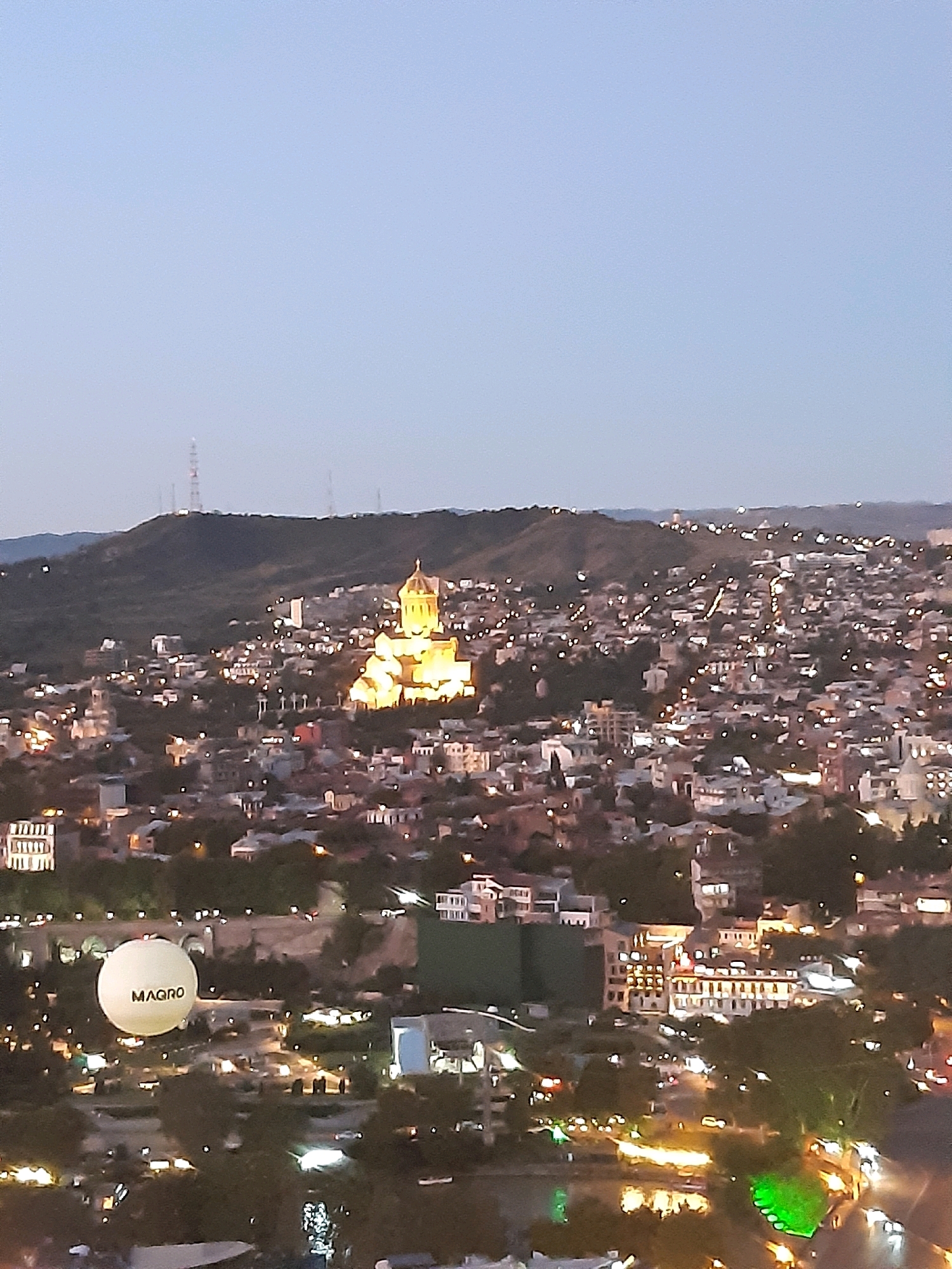 The image shows an aerial view of the city of Tbilisi, Georgia, at dusk. In the center of the frame, the illuminated Holy Trinity Cathedral of Tbilisi (Sameba Cathedral) stands prominently on a hill, its golden dome shining brightly against the darkening sky. Below and around the cathedral, the cityscape is dotted with numerous lights from buildings and streetlamps, creating a warm glow. In the foreground, a large white hot air balloon with the word "MAGRO" printed on it is visible, floating above the city. The background features rolling hills and a distant transmission tower. The overall impression is of a vibrant city at twilight, with a mix of historic and modern architecture.