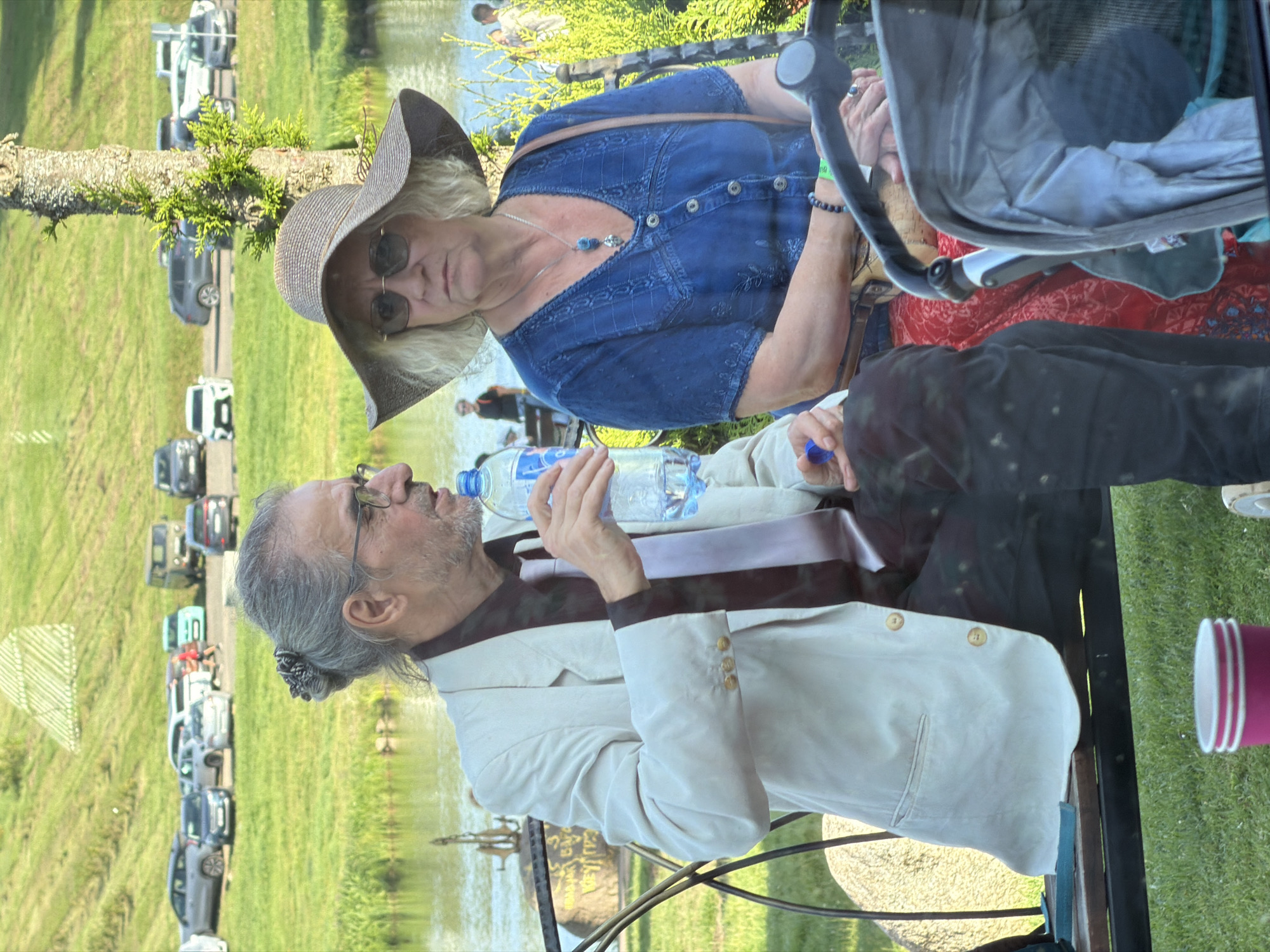 The image shows an outdoor scene with two people, a man and a woman, seated on a bench. The man is wearing a light-colored jacket, a dark shirt, and a light-colored tie. He has glasses and his hair is tied back. He is holding a bottle of water. The woman is wearing a wide-brimmed hat, sunglasses, and a blue top. She has a necklace and bracelets on. They are positioned in front of a body of water and a grassy slope with parked cars in the background. A baby stroller is partially visible in the foreground. The setting appears to be a park or recreational area. The location is identified as Litvyenski Sel'ski Sovet, Belarus.