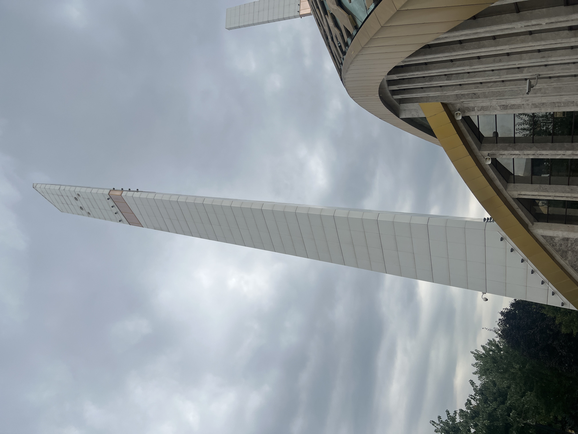 The image shows a low-angle view of the Argun Central Mosque in Argun, Russia. The mosque features a tall, white, slender minaret that tapers towards the top, with a band of copper-colored material partway up. To the right of the minaret, the distinctive arched roof of the mosque is visible, adorned with copper-colored cladding and vertical concrete supports. Another, shorter minaret is partially visible behind the main structure. The sky is overcast with gray clouds. Green trees can be seen to the left of the minaret. Security cameras are mounted on the building.