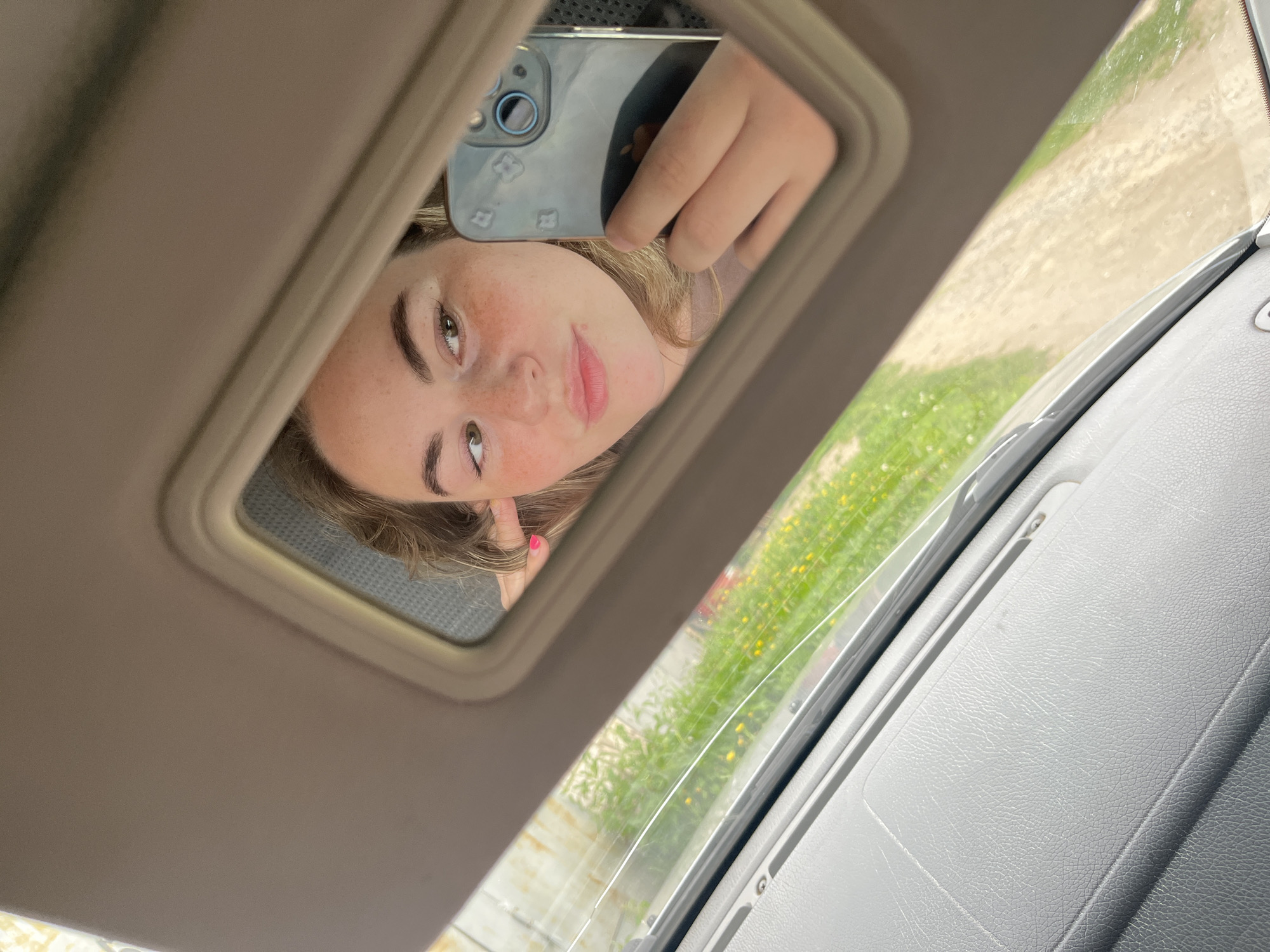 A person is seen from the chest up, reflected in a car's beige overhead vanity mirror, positioned in the city of Chaikovsky, Russia. The individual, appearing to be female with light brown hair and visible freckles on her nose and cheeks, holds a smartphone in her right hand to capture the reflection. Her left hand, with brightly painted pink fingernails, is casually positioned near her left ear. Her gaze is directed towards her right (our left), and her lips are slightly puckered. The smartphone, identifiable as an iPhone by its triple camera array and partially visible Apple logo through a clear, subtly patterned case, is prominent in the reflection. Below the mirror, the gray, textured dashboard and a portion of the windshield are visible, revealing an outdoor scene. This scene includes a dirt or gravel path, flanked by vibrant green grass interspersed with small yellow flowers, under bright daylight conditions. A light-colored structure or fence line is faintly visible in the upper left background of the outdoor view.