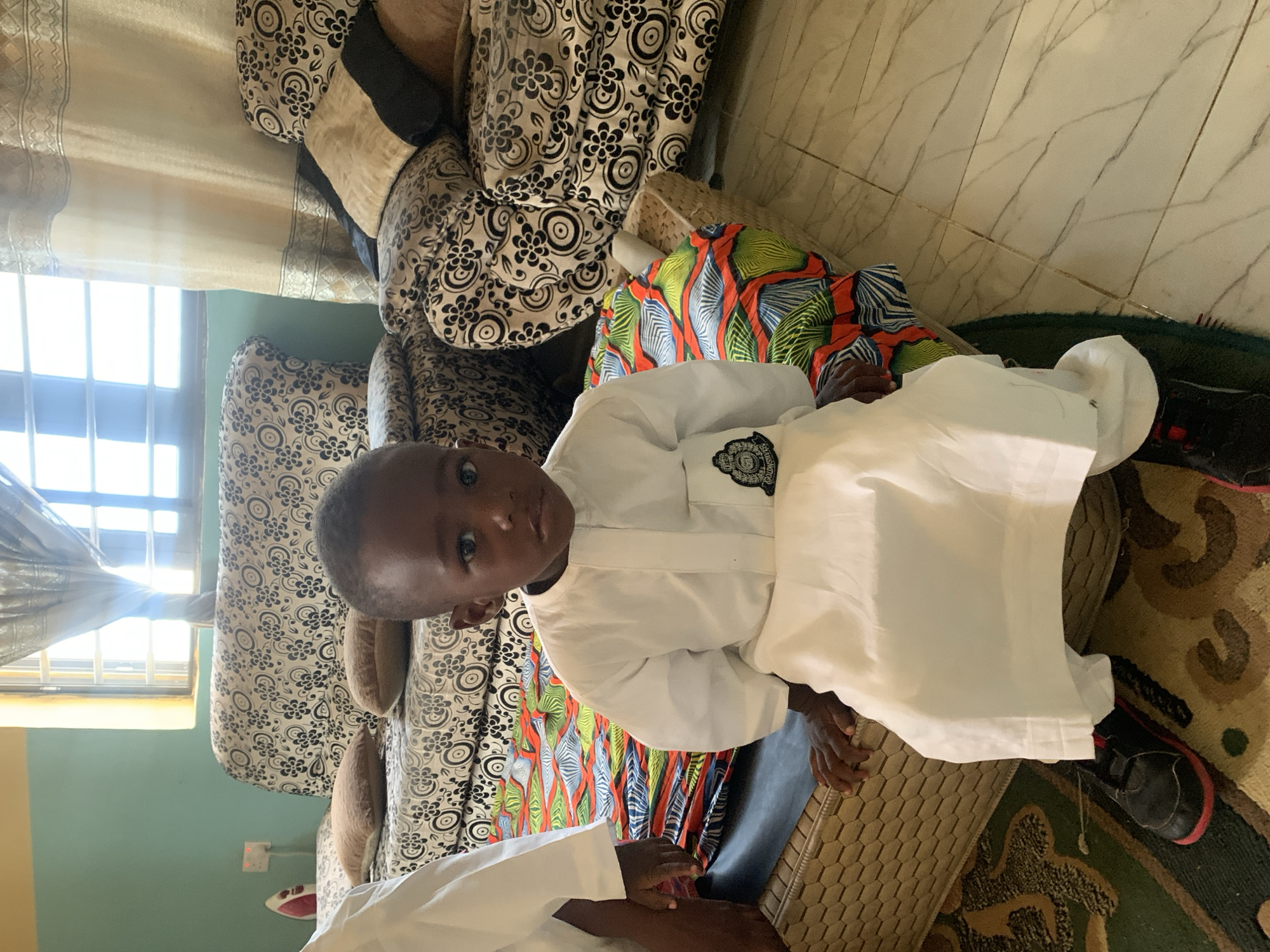 A young child wearing a white, long-sleeved garment is sitting on a woven surface. The child has dark skin and is looking at the camera. To the left, there is a patterned sofa with throw pillows. Behind the child, there is a colorful patterned cloth and part of a green rug. The background includes a window with curtains, a wall painted in green, and an electrical outlet. Part of a person wearing a white garment is visible in the foreground. The location is Gura Top, Nigeria.