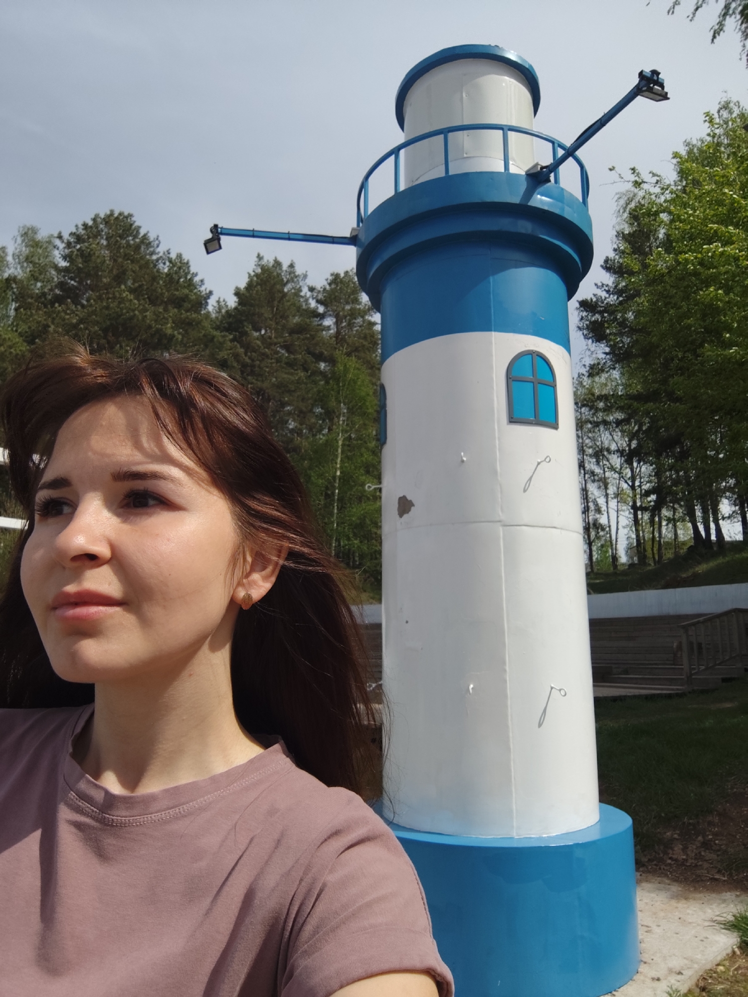 A young woman with brown hair and a mauve t-shirt is standing outdoors in Chaikovsky, Russia, posing in front of a blue and white lighthouse replica. The lighthouse has a blue base, a white middle section with a small blue-framed window, a blue upper section, and a white top. It is equipped with a lighting fixture on a pole extending outward. The background includes green trees, a clear sky, and part of a wooden structure, possibly stairs. The setting is likely a park or recreational area within the city.