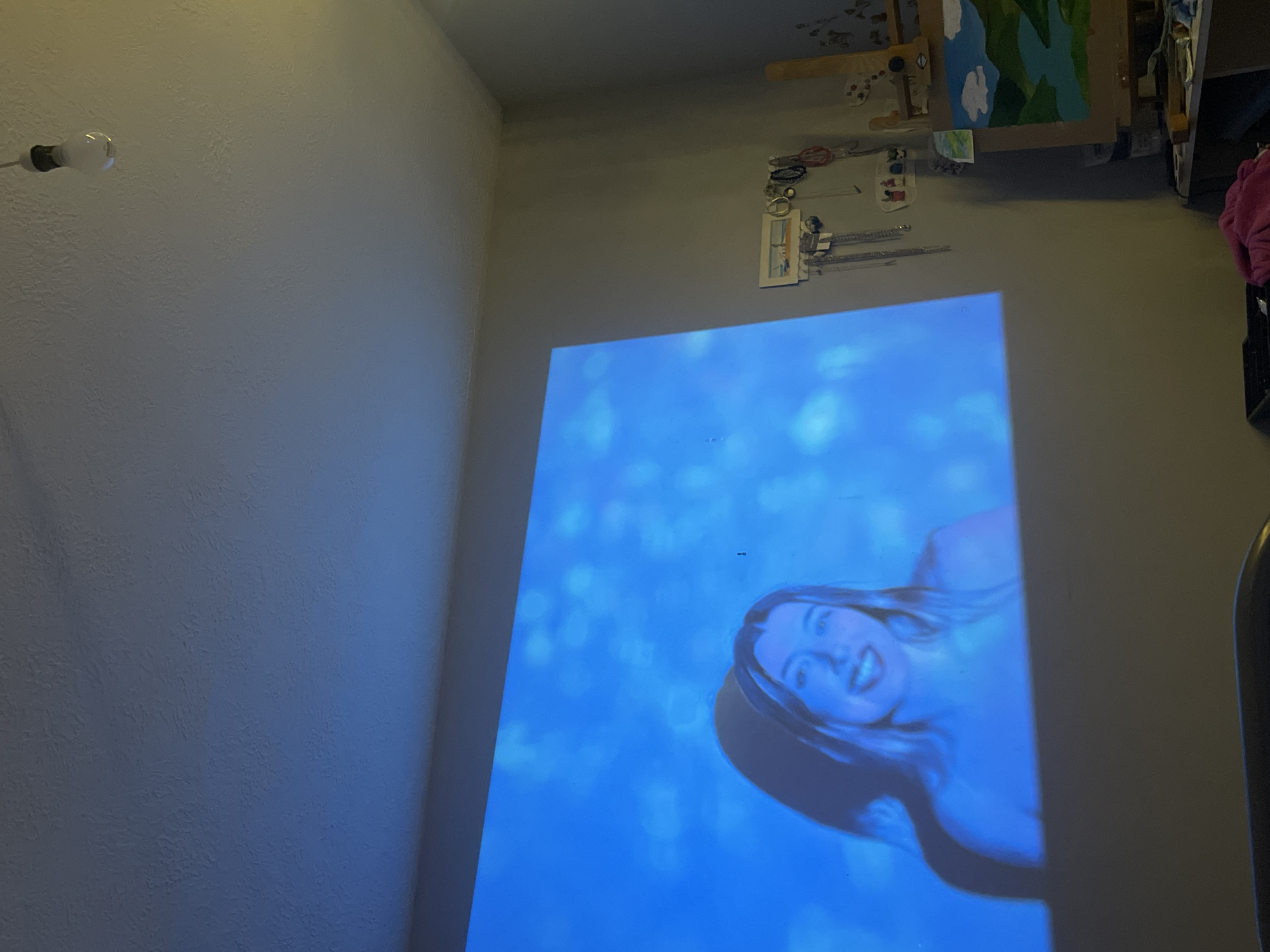A projected image of a woman's face, seemingly surrounded by water, illuminates a wall. The environment appears to be an interior space with household objects and what looks like a painting easel, partly visible on the upper right. A lightbulb is also visible, though it's uncertain if it's illuminated. Without further information, specifying a location within Maidstone, United Kingdom is not possible.