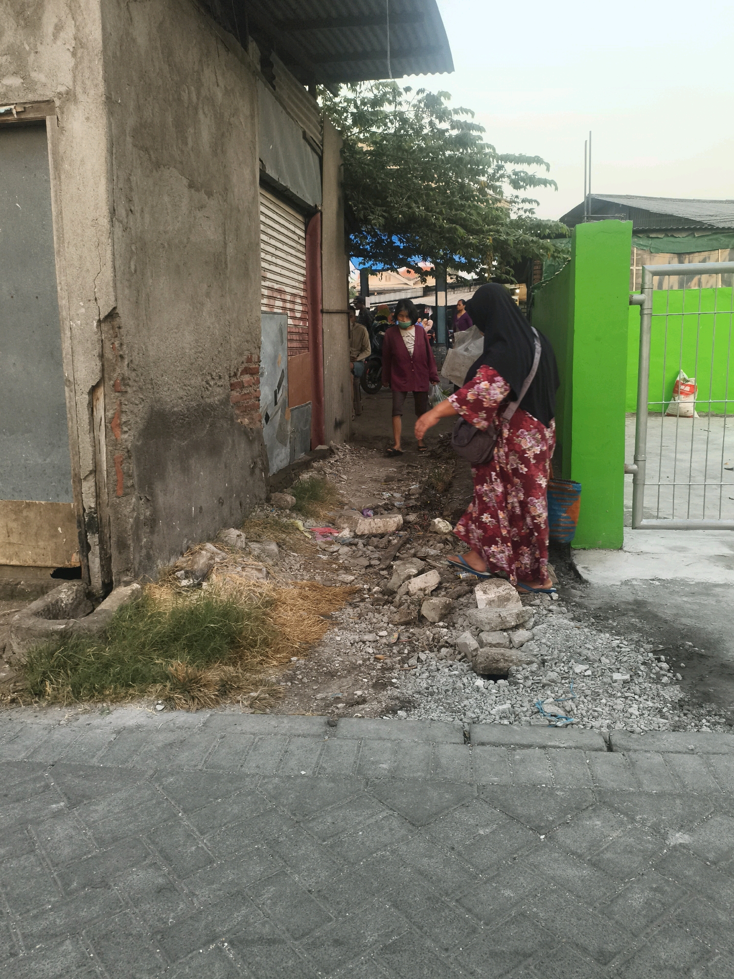 The image depicts a street scene in Surabaya, Indonesia. In the foreground, a woman wearing a black hijab and a floral-patterned dress walks to the right. She has a brown crossbody bag. Her feet are clad in blue flip-flops. The ground beneath her is a mixture of grey pavement and rough, rocky terrain littered with debris. To her left, there is a building with a rough concrete exterior, showing exposed brickwork in some areas. A metal roller shutter door is visible on this building. Further into the background, a bright green wall and a metal fence are prominent. Behind the green wall, a white sack is visible. Trees with green foliage are present in the middle ground. Other individuals are faintly visible in the background, some appearing to be walking or standing near the buildings. The overall environment appears to be an urban setting with elements of construction or disrepair. The sky is overcast.
