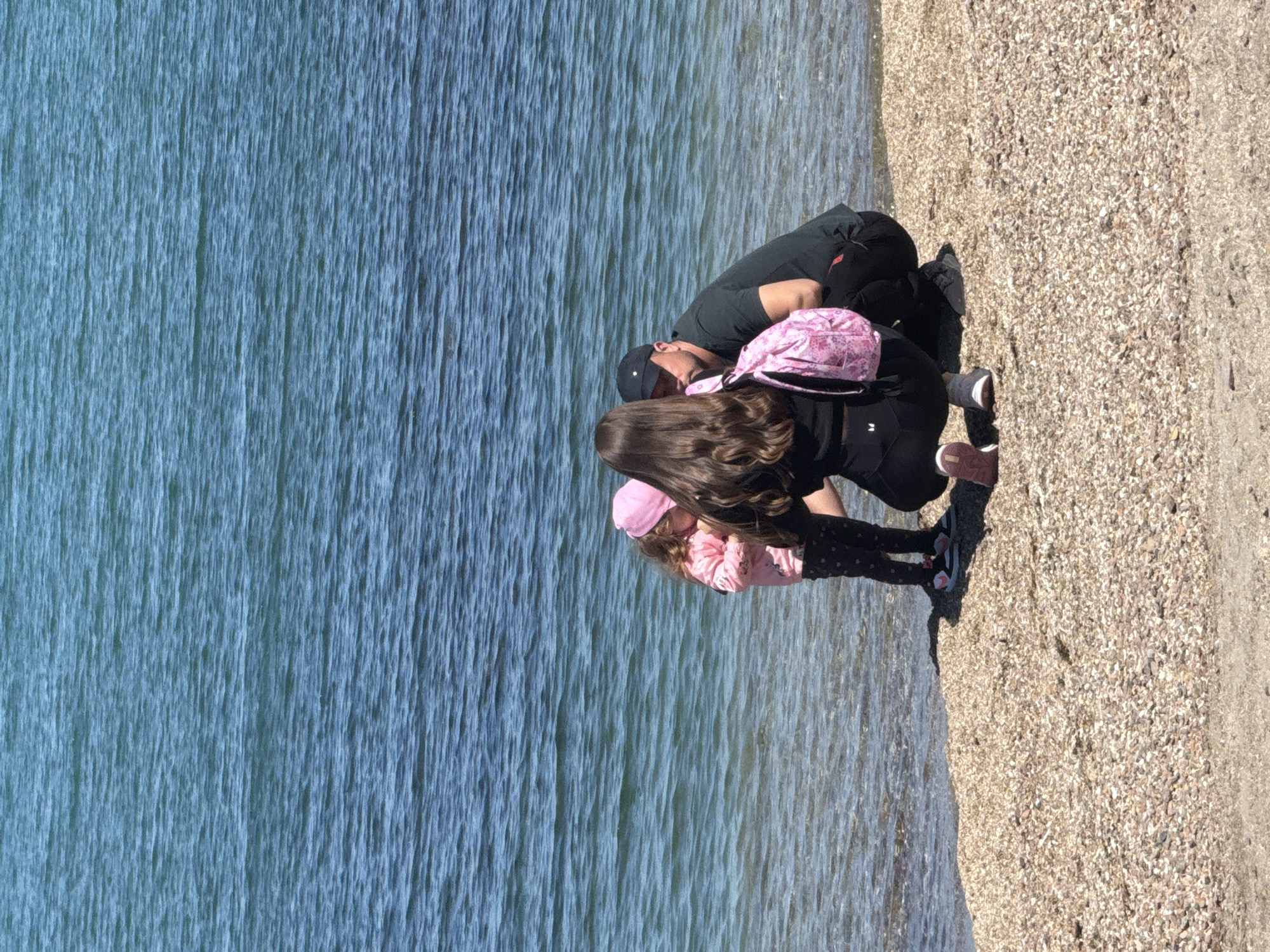 The image shows three people on a beach next to the Black Sea, likely in Burgas, Bulgaria. The beach consists of small pebbles. The water is calm, reflecting light in vertical streaks. An adult male is squatting with his head bowed, facing a young girl. Behind him, another female is also squatting. The young girl is wearing a pink shirt, a pink hat and patterned leggings. The female has long brown hair and is wearing a black jacket and a pink backpack. The man is wearing a black t-shirt and a baseball cap. They appear to be looking at something on the ground. Shadows are cast by the figures, indicating bright sunlight.