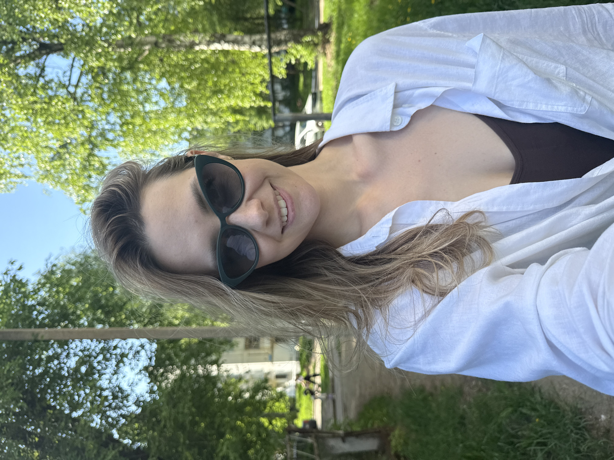A woman with light brown hair and wearing sunglasses smiles at the camera. She is wearing a white, button-down shirt over a brown top. The background shows a green, grassy area with trees and a building in the distance. Based on the location in the city of Chaikovsky, Russia, the environment is likely a park or residential area. The image appears to be a selfie taken outdoors during daylight.