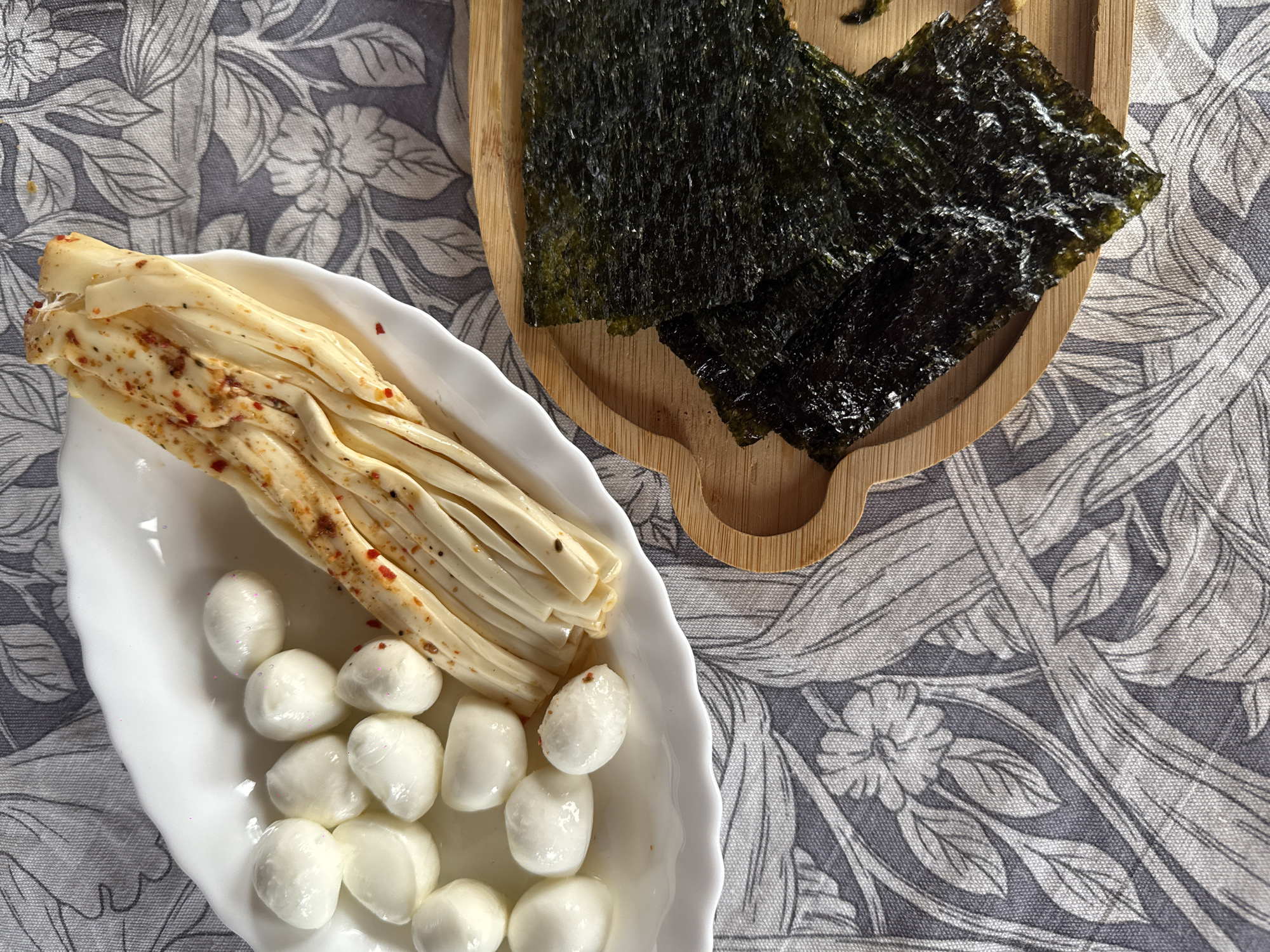 An overhead, static shot captures a variety of food items arranged on a patterned textile surface. The surface displays an intricate grey and light grey floral and foliate design.
In the upper left, a white, scalloped-edge plate holds two distinct food preparations. On the left side of the plate are approximately twelve small, white, roughly spherical food items, consistent with mini mozzarella balls (bocconcini or ciliegine). Several of these items exhibit faint pinkish-purple specks on their surface and are partially submerged in a clear liquid. To their right, several strands of yellowish, flat, and pliable strips are arranged, coated with visible reddish-orange chili flakes and dark specks, consistent with seasoned string cheese, possibly Chechil.
In the lower right, a curved wooden serving board, light brown with visible grain, holds multiple rectangular sheets of dark green, crinkled, and glossy roasted seaweed snacks (nori).
No individuals are present in the image. The setting appears to be an indoor dining or snacking arrangement. No geographic indicators are present to suggest the location is the city of Фоки, Russia.