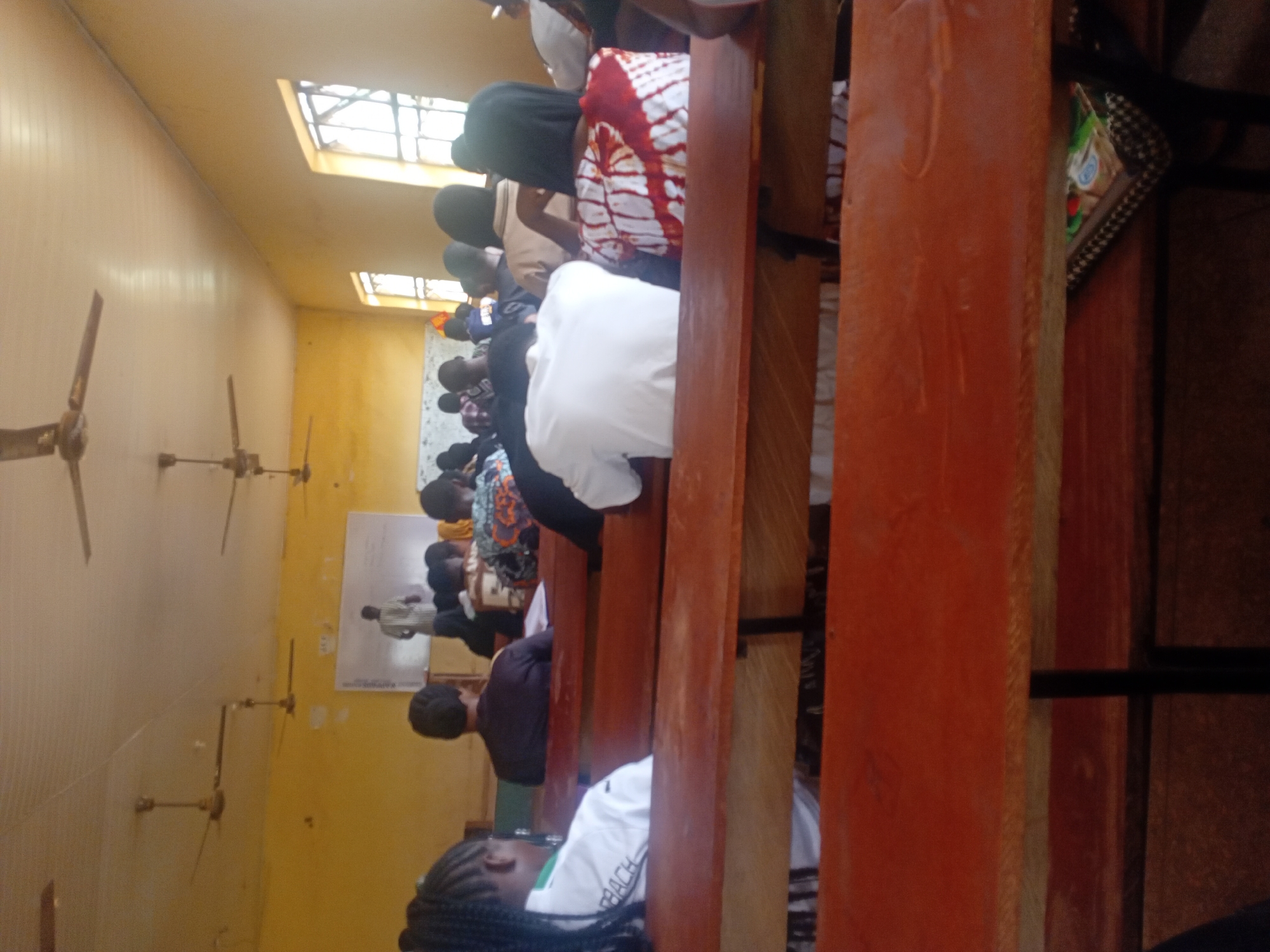 The image shows a gathering of people seated on benches inside a room. The room has yellow walls, and there are several ceiling fans. A poster is visible on one wall. Natural light enters through windows near the ceiling. The people appear to be focused on something ahead, possibly a presentation or speaker. The location is in the city of Unknown, Unknown.