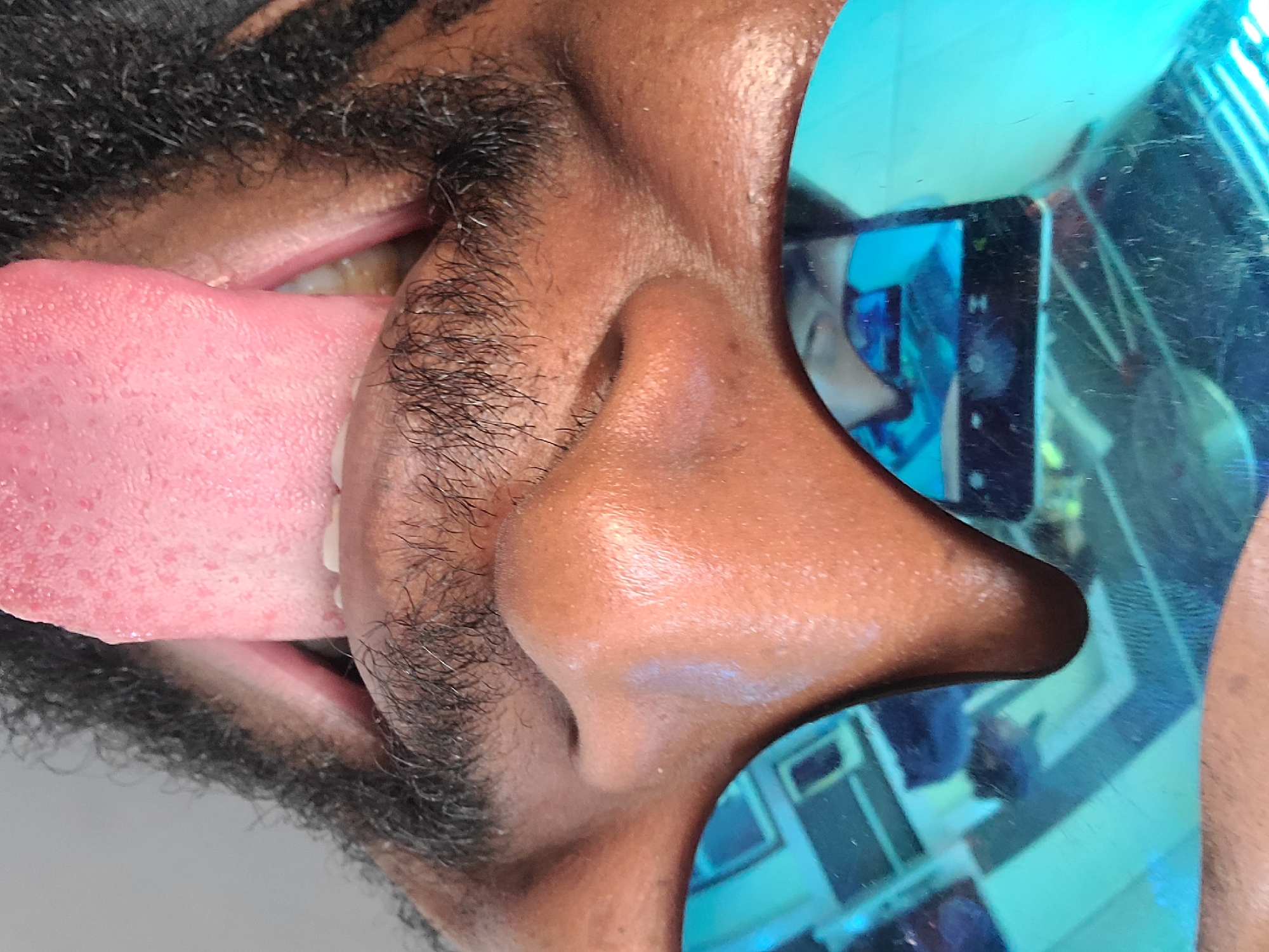 This close-up portrait, rotated ninety degrees clockwise, features a person from Jos, Nigeria, captured from a low angle. The subject has dark skin, a prominent nose, and a thick, curly black beard and mustache. Their mouth is wide open, with a large, pink tongue extended forward, displaying visible papillae. A few slightly discolored upper teeth are visible behind the tongue.
The person is wearing bright blue, highly reflective sunglasses on their right eye (which appears on the left side of the image due to the rotation). The large, oval lens of the sunglasses reflects an indoor scene. The reflection shows what appears to be a room with light blue elements, possibly geometric patterns on a wall or floor, and a dark screen or monitor in the distance. A faint letter "H" is discernible on the reflected screen. The blurry reflection also includes indistinct shapes that suggest furniture or other people, indicating an interior setting. The extended tongue gives the subject a playful or whimsical expression.